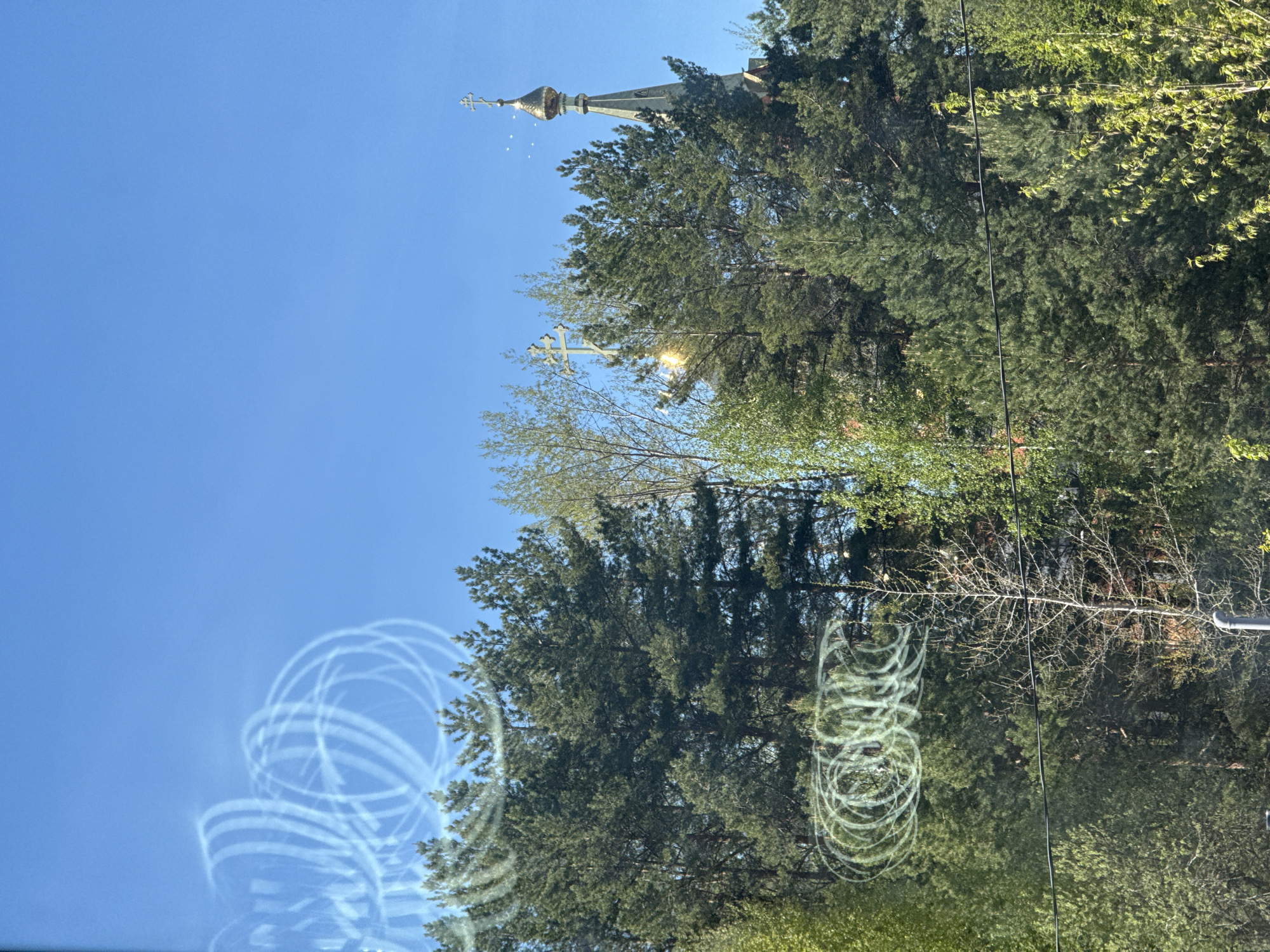 The image shows a view of a Russian Orthodox church spire peaking above trees. The sky is blue. There are swirls on the glass that appear to be reflections. Overhead wires can be seen. This is likely in the city of Unknown, Unknown.