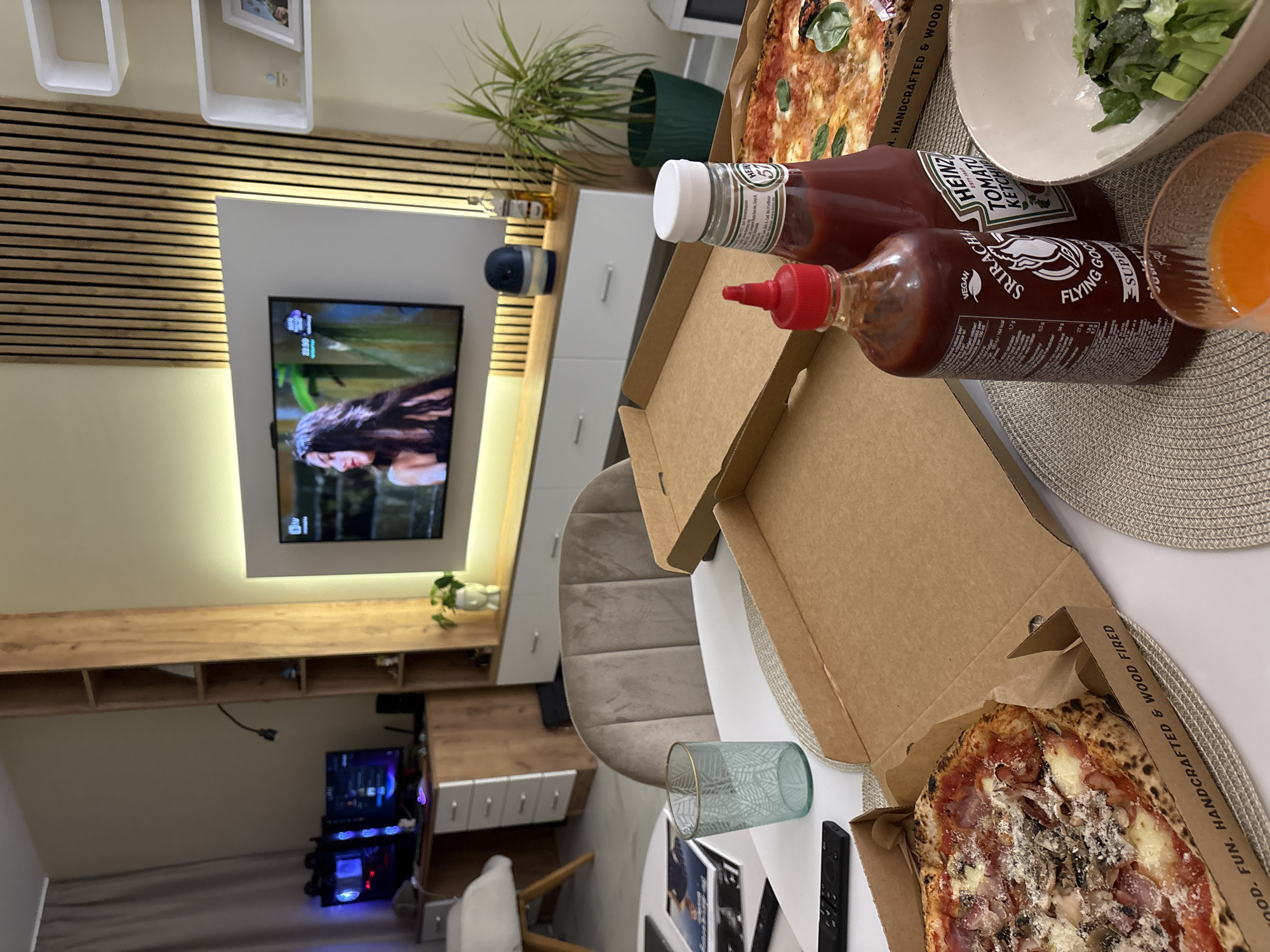 The scene shows a partially eaten meal of pizza, salad, juice, ketchup, and Sriracha sauce on a round table with a placemat. The pizza is in two open cardboard boxes labeled "HANDCRAFTED & WOOD FIRED" and "FUN." The sauces are identified as Heinz Ketchup and Flying Goose Sriracha. Behind the table is a living room with a TV displaying an image of a person. The location of the image is a room in Sofia, Bulgaria.