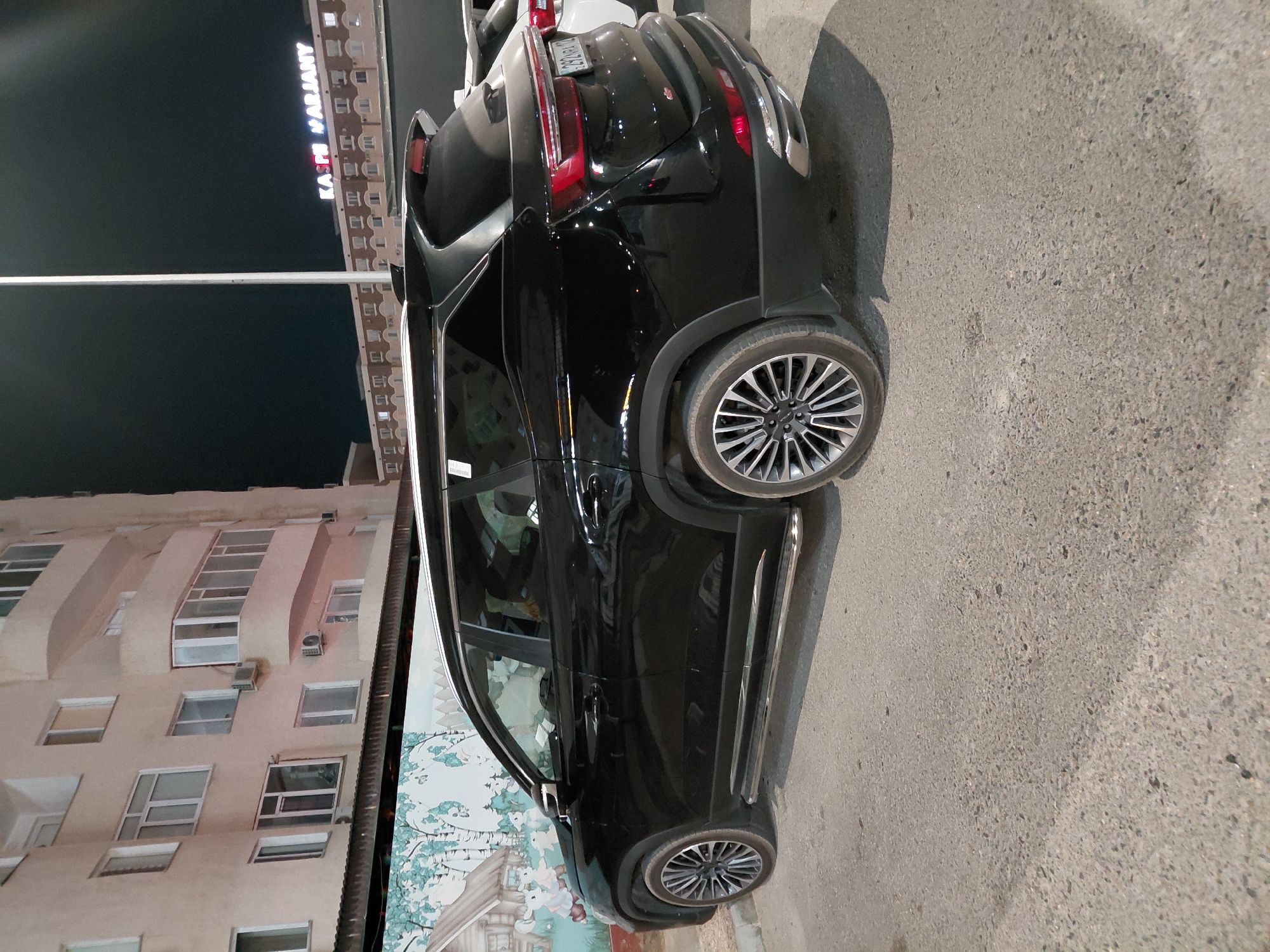 A black SUV is parked on a paved surface in front of a multi-story building in Aktau, Kazakhstan. The building has cream-colored walls, numerous windows, and balconies. A mural featuring trees and small houses is visible on the lower part of the building's facade. The SUV has chrome accents on its side and rear bumper. The license plate is partially visible and appears to be in Cyrillic script. In the background, a tall light pole and a building with illuminated signage that reads "KASPIY MARJANY" are visible against a dark sky. Another white vehicle is parked behind the black SUV. The scene appears to be at night or in low light conditions.