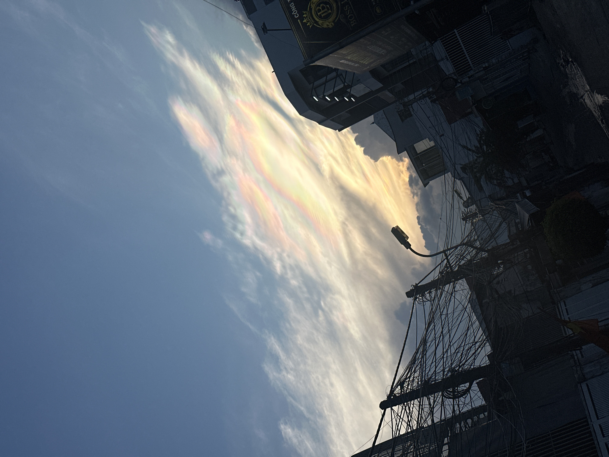 The image captures a view in Bac Nha Trang Ward, Vietnam. The sky dominates the upper portion of the scene, with elongated, iridescent clouds stretching across a blue expanse. These clouds display a range of colors, including pink, yellow, and white.
In the lower right of the image, the urban landscape is visible, featuring buildings, power lines, and a street lamp. The dense network of power lines creates a web-like pattern against the sky. A building with a sign that says "BOSS" is also present. The perspective suggests the image was taken from a low angle, looking upwards.