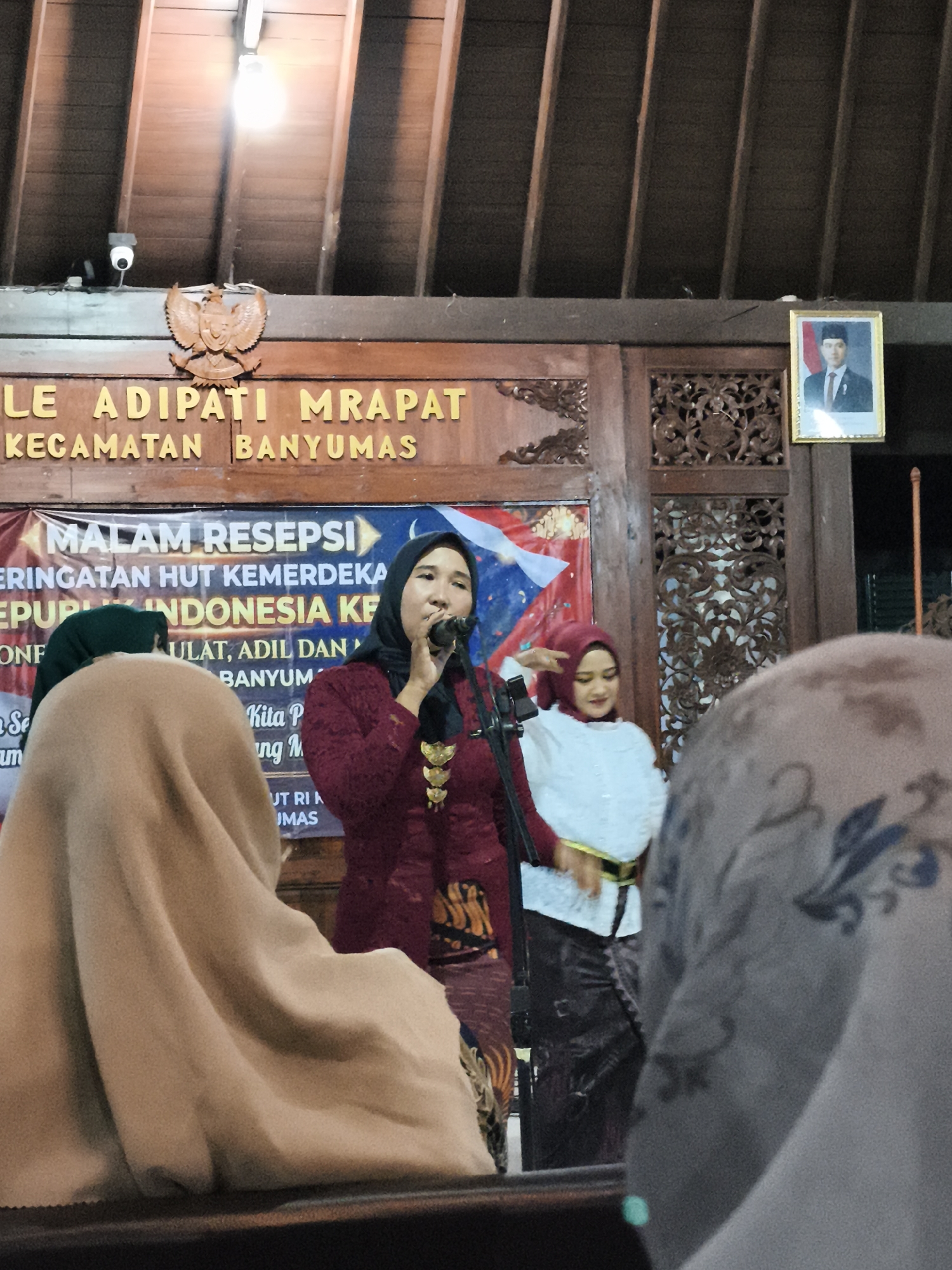 In an indoor setting, likely a community hall or local government office in Banyumas, Indonesia, a woman wearing a maroon hijab and a matching top is speaking into a microphone. She is positioned in the center of the frame and appears to be addressing an audience. Behind her, a banner with Indonesian text, including "MALAM RESEPSI" and "REPUBLIK INDONESIA KE," is visible. To her left and right, other individuals are present, some partially obscured by foreground elements. One woman to her right wears a white top and a red hijab, and another person in the foreground on the left is covered by a tan hijab. The background features wooden architectural elements, including carved panels, and a framed portrait of a man in formal attire is hung on the wall. Overhead lighting illuminates the scene.