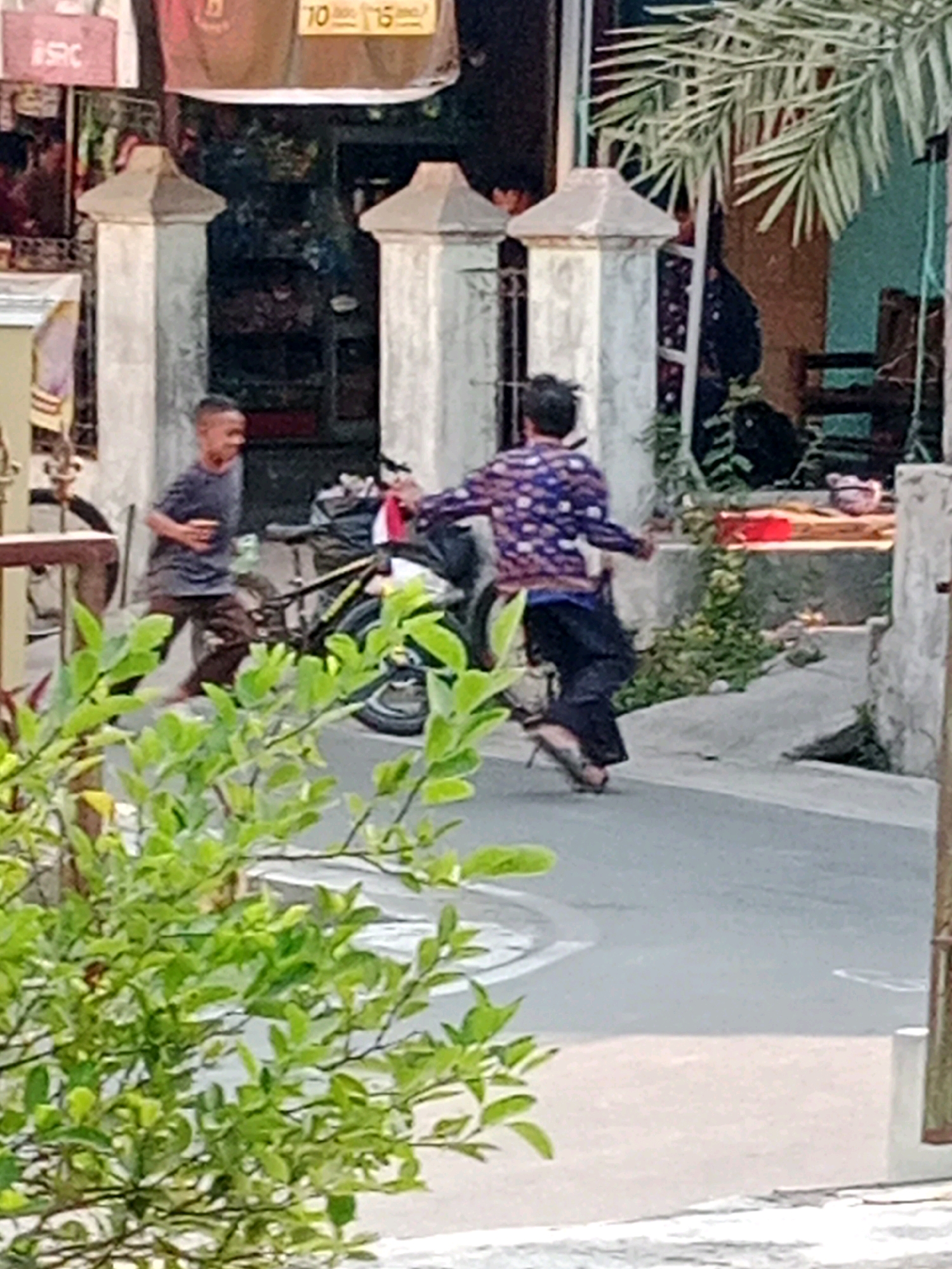In Waru, Indonesia, two young boys are depicted on a street. One boy, wearing a dark t-shirt and brown pants, is captured mid-stride, facing left with his body slightly turned. He appears to be moving dynamically, possibly in the middle of a race or playful chase. Adjacent to him, a bicycle with a black frame and yellow accents is parked, its rear wheel partially obscured by the boy. To the right, another boy, dressed in a patterned purple and white long-sleeved shirt and dark pants, is in motion, his back mostly visible as he runs towards the right side of the frame. He seems to be actively participating in the same activity. The background reveals a setting with a building featuring decorative pillars and a hint of foliage. The scene is observed through a screen of vibrant green leaves in the foreground, suggesting the vantage point is from across a street or a garden. The street surface is paved, with visible white markings.