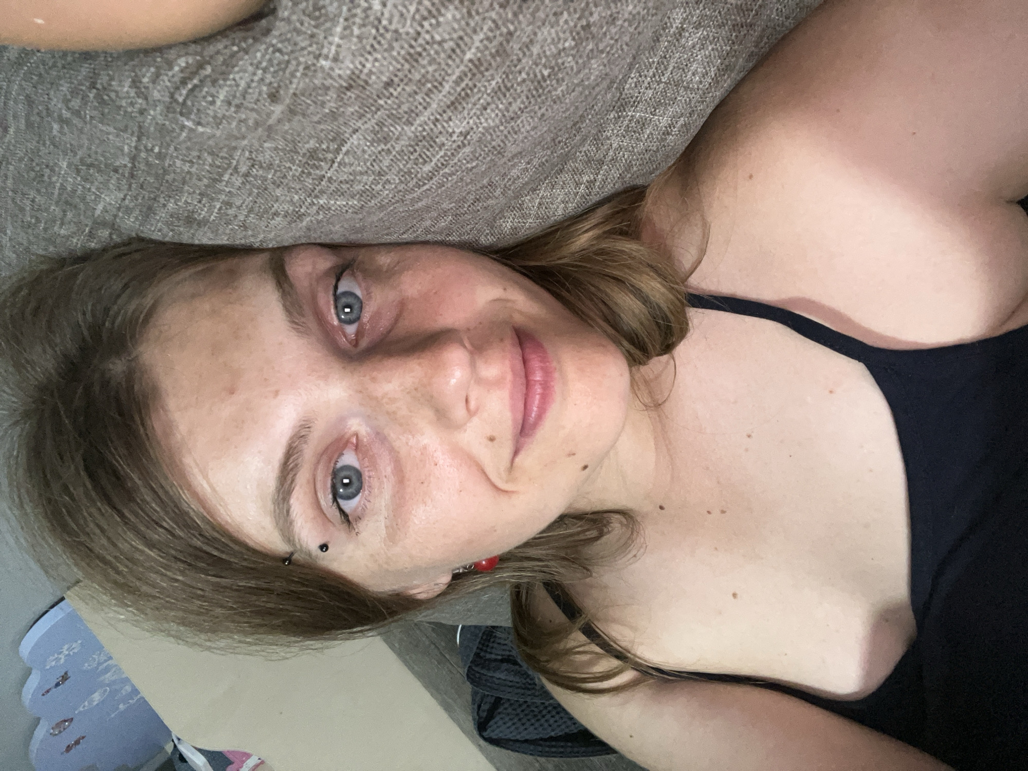 A young adult with light brown hair, blue eyes, and freckles is lying on a couch. They are wearing a black tank top and have a black eyebrow piercing. The person is looking up and slightly to the left, with their head resting on a grey textured cushion. The background shows a blurred interior scene, including what appears to be a light blue decorative object. The overall impression is a casual, relaxed setting. The location is not discernible from the image.
