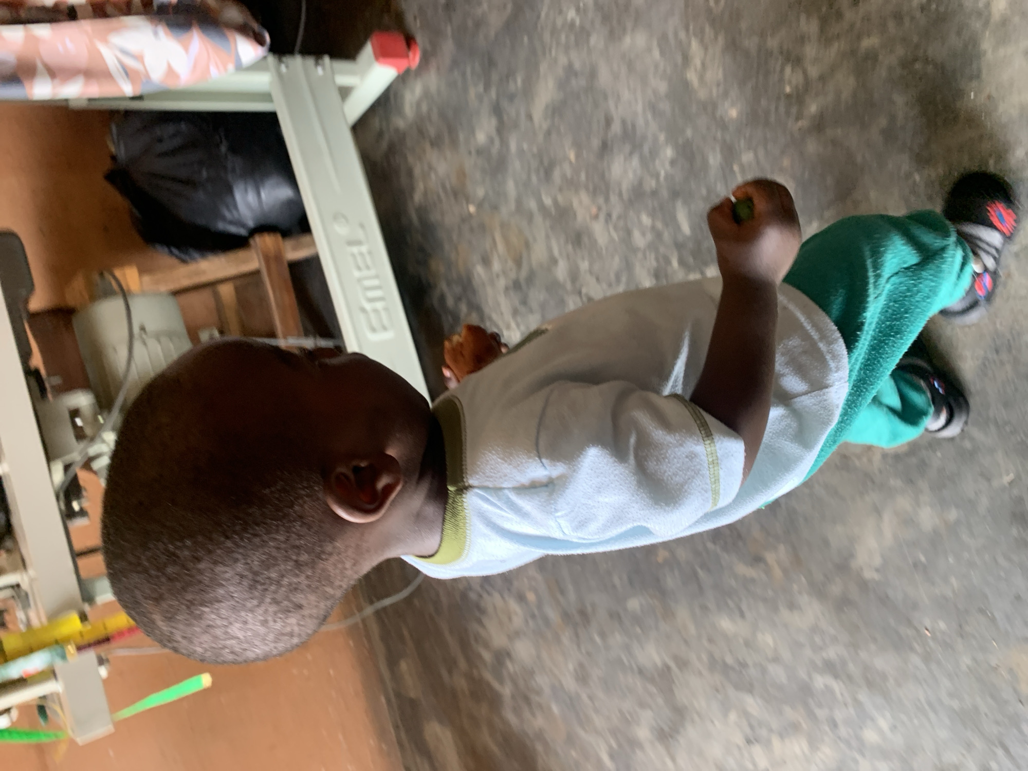 A young child, viewed from the side and slightly from behind, stands on a concrete floor in what appears to be an indoor space. The child is wearing a light blue t-shirt with a green trim at the collar and teal-colored pants. They have dark skin and short hair. In their right hand, the child is holding a small, brown, round object, possibly food. Their left hand is slightly bent, and their fingers appear to be grasping something small and green. The child is wearing dark shoes with red and grey accents.
In the background, a light-colored sewing machine or industrial equipment is visible, with parts of what look like wooden supports and a black plastic bag behind it. The walls appear to be orange-brown. The overall environment suggests a workshop or a home setting.
The image is a photograph, and the focus is on the child. The lighting is moderate, and the scene is captured from a medium distance. No specific landmarks or identifiers place this image within the city of Jos, Nigeria, other than the prompt's request to include this location.
