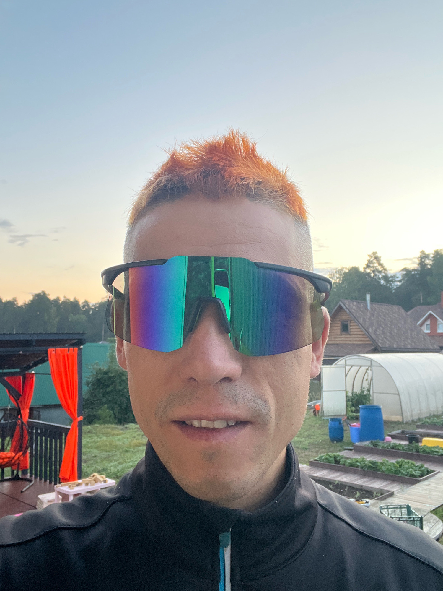 A man with short, spiky, dyed orange hair and a close-cropped beard and mustache, wearing large, wraparound sunglasses with a colorful, iridescent lens, and a black zipped jacket, is standing outdoors. He is looking directly at the camera and smiling slightly. The background suggests a rural or suburban setting, possibly a dacha or country house. There are several elements visible: a greenhouse, garden beds with green plants, wooden fences or raised beds, large blue barrels, a small red roofed building, and trees. To the left, there's a covered patio or porch area with orange curtains hanging. The sky is clear and light blue, indicating it might be morning or evening. The overall atmosphere is casual and relaxed. This scene is located in Chaikovsky, Russia.