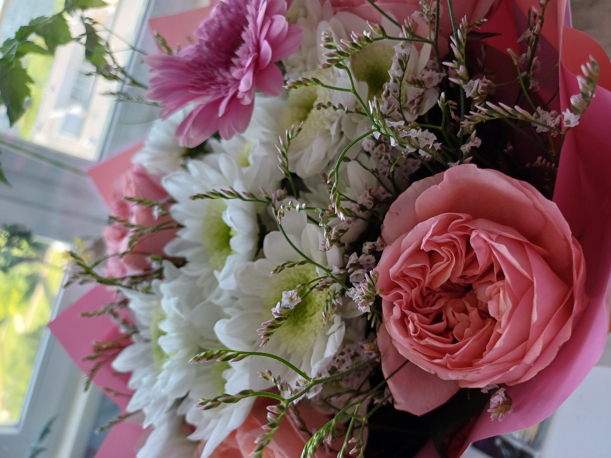 A close-up, eye-level, vertical shot shows a bouquet of flowers in pink and white hues, positioned in front of a window with an outdoor view. The bouquet is arranged with a large, tightly petaled pink rose in the foreground on the left, surrounded by numerous white daisy-like flowers with yellow centers. A bright pink gerbera daisy is visible on the right side of the bouquet, partially obscuring some of the white flowers. Delicate green stems with small white and pink buds are interspersed throughout the arrangement, adding texture and detail. The bouquet is wrapped in pink paper, with some of the paper extending upwards and to the left, creating a backdrop. The window behind the bouquet reveals a blurred view of buildings and greenery outside, suggesting an urban setting. The lighting is natural, coming from the window, which illuminates the flowers and creates soft shadows.