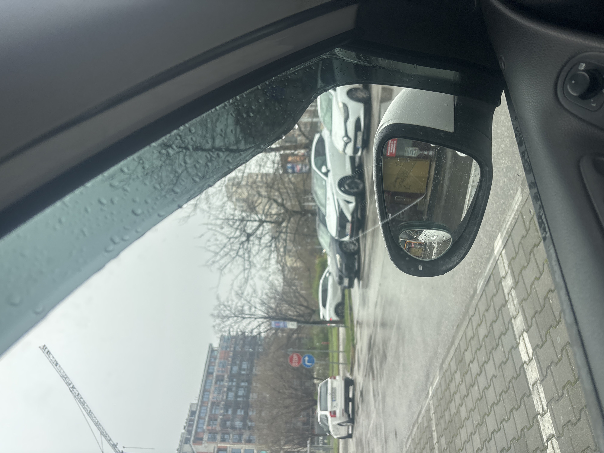 The image depicts a view from inside a car, looking out towards a street scene in Plovdiv, Bulgaria. The view is partially obscured by a rain-streaked car window and the car's side mirror. The mirror is cracked. Reflected in the side mirror is a section of the street, including a row of parked cars and a building. Outside the car window, a construction crane is visible against a cloudy sky. In the distance, an apartment building is visible, along with bare trees and street signs. The pavement is wet, suggesting it is raining. The street appears to be made of grey paving blocks.