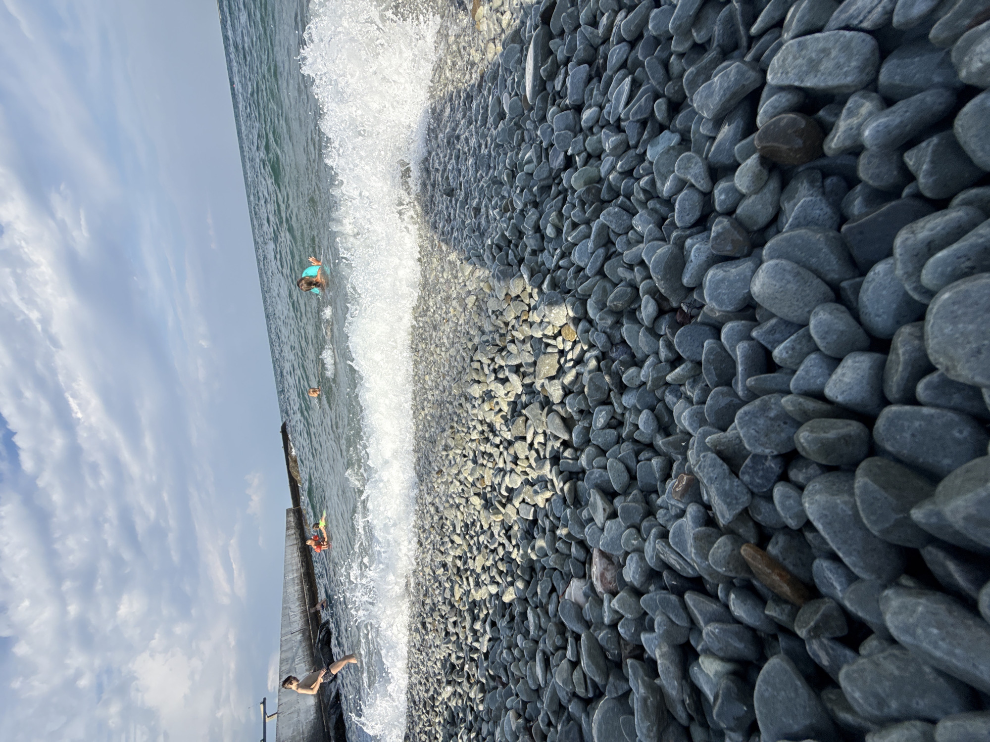 In Partenit, Ukraine, a body of water laps against a pebble beach under a cloudy sky. A wave crashes onto the shore, creating a foamy white crest. In the distance, several people are in the water. On the left side of the frame, a boy stands near a concrete structure that extends into the water. The beach is covered in smooth, gray pebbles of varying sizes. The scene is an outdoor, daytime shot.