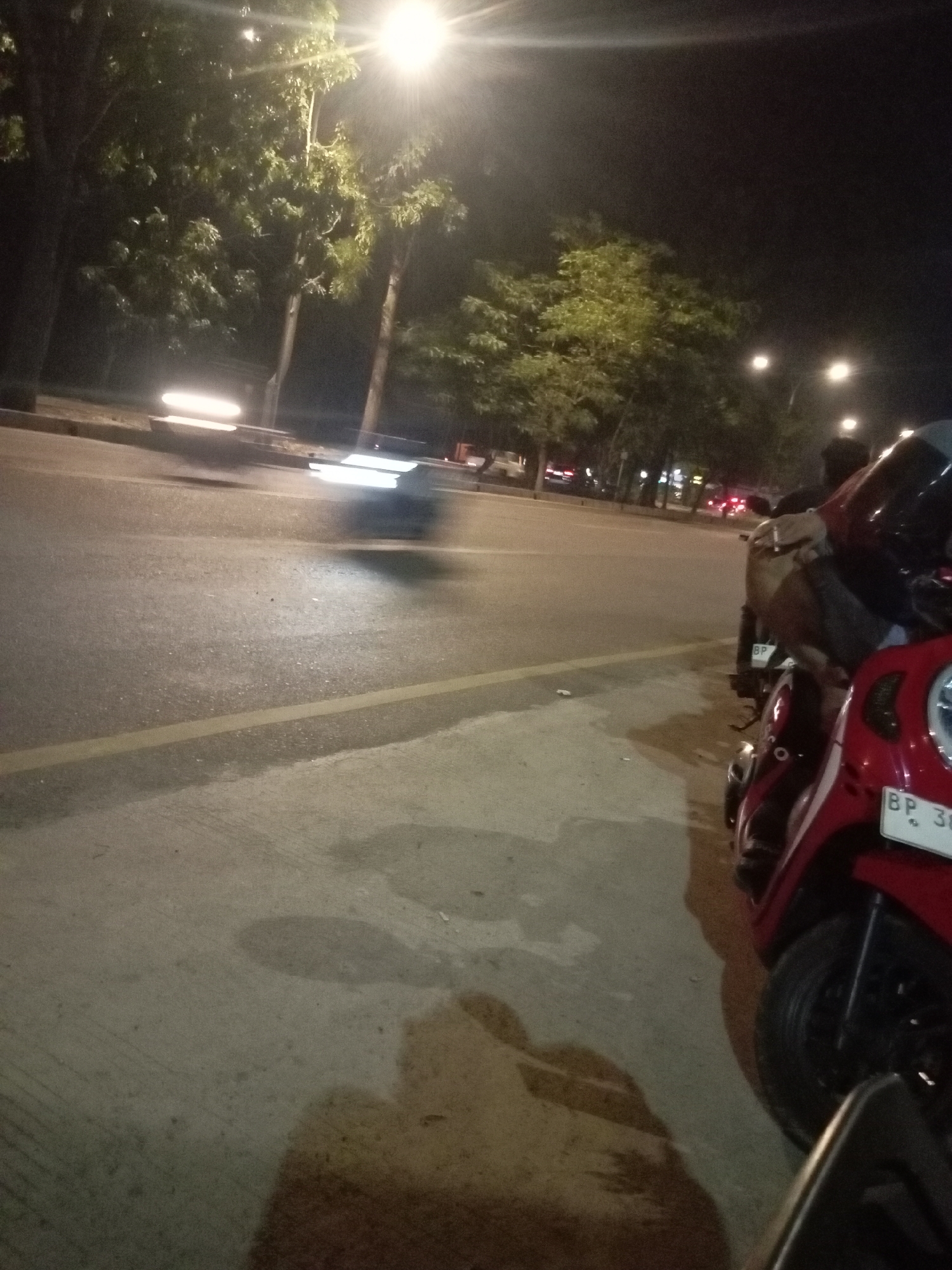 The image depicts a street scene at night in Batam, Indonesia. A street lamp illuminates the area, casting light on a road and a sidewalk. On the right side of the frame, two red scooters are parked. The scooter in the foreground has a license plate that reads "BP 38". The road shows signs of traffic, with blurred lights suggesting moving vehicles. Trees line the street in the background. The overall atmosphere is dark, typical of a nocturnal urban environment.