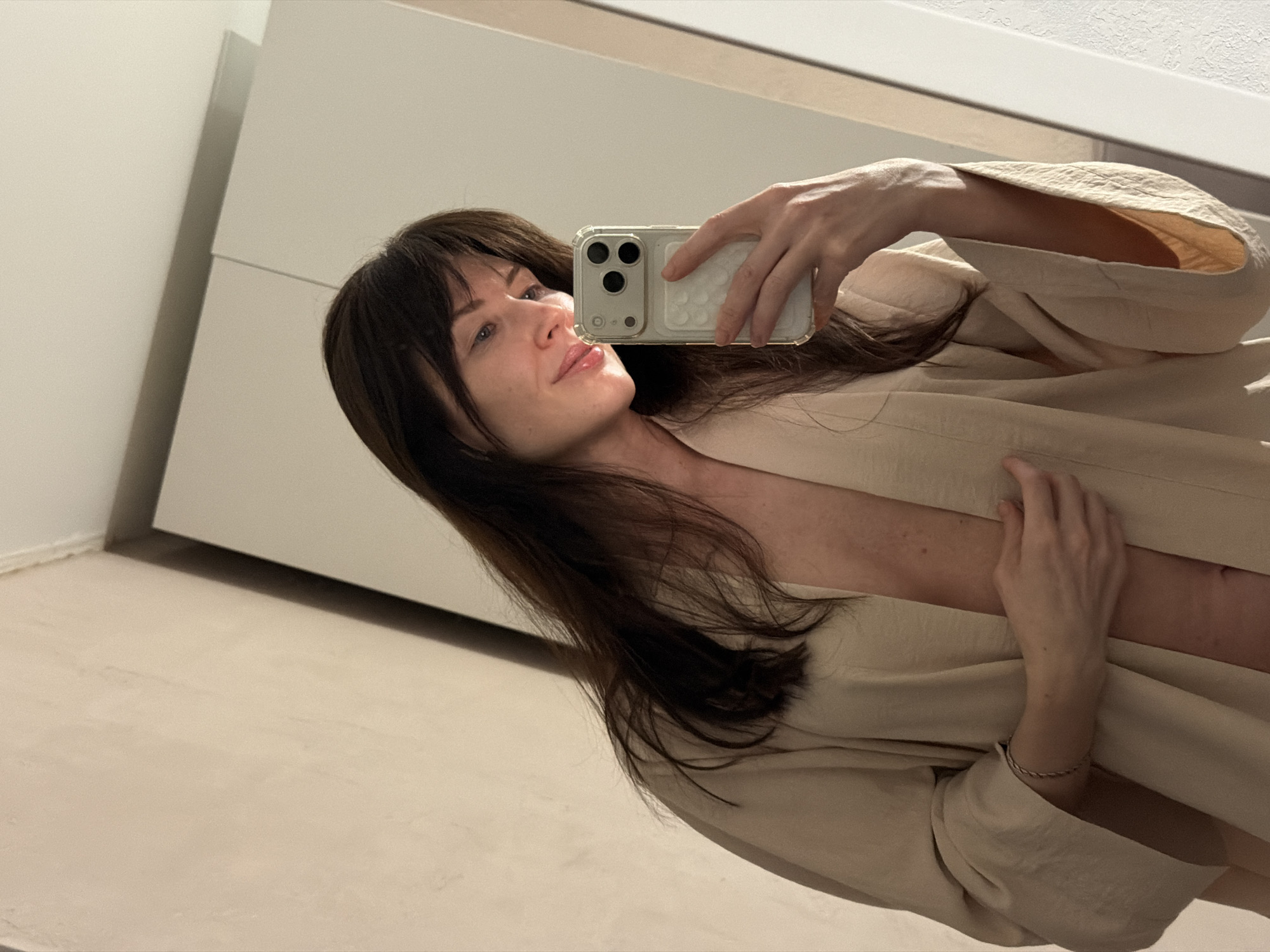 A woman with long, dark hair and bangs is captured in a mirror selfie, holding a smartphone with a clear case and triple camera system. She is looking upwards and slightly to her left, displaying a gentle smile. Her fair skin is visible against the light-colored, loose-fitting garment or robe she is wearing, which appears to be beige or off-white. Her left arm is raised to hold the phone, and a delicate bracelet is visible on her right wrist.
The setting appears to be indoors, likely an apartment or hotel room in Perm, Russia. The background consists of minimalist, light-colored walls or cabinetry and a light-toned floor, suggesting a modern interior. The lighting is soft, illuminating her face and the immediate surroundings. The image is tilted, giving a dynamic angle to the mirror reflection. No specific time of day or weather conditions are discernible, but the interior lighting is even. There is no visible text in the image. The overall mood is calm and reflective.