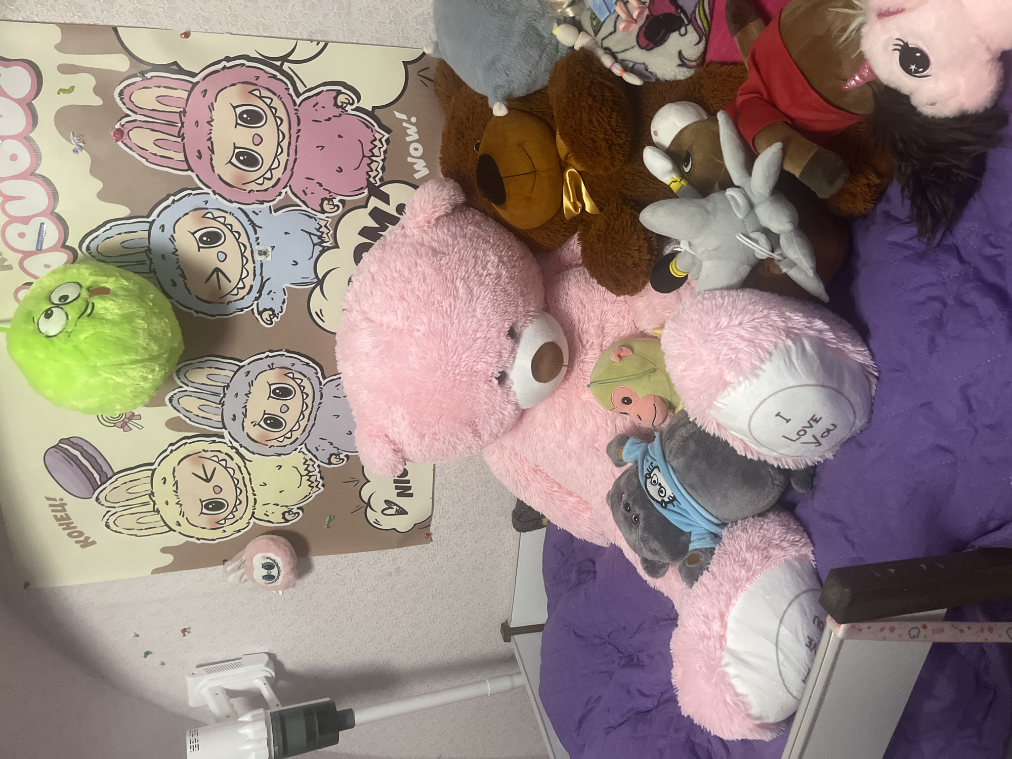 The image displays a collection of stuffed animals on a purple surface, likely a bed. A large pink teddy bear is prominently featured in the center, with a brown teddy bear to its right. Several other stuffed animals, including a grey cat, a green monkey, and various character-themed plush toys, are scattered around. A poster with cartoon characters and text is visible on the wall behind the stuffed animals. A white vacuum cleaner is mounted on the wall to the left. The setting appears to be an indoor environment, possibly a child's bedroom. There is no visible indication of the specific location within Vladimirovka, Russia.