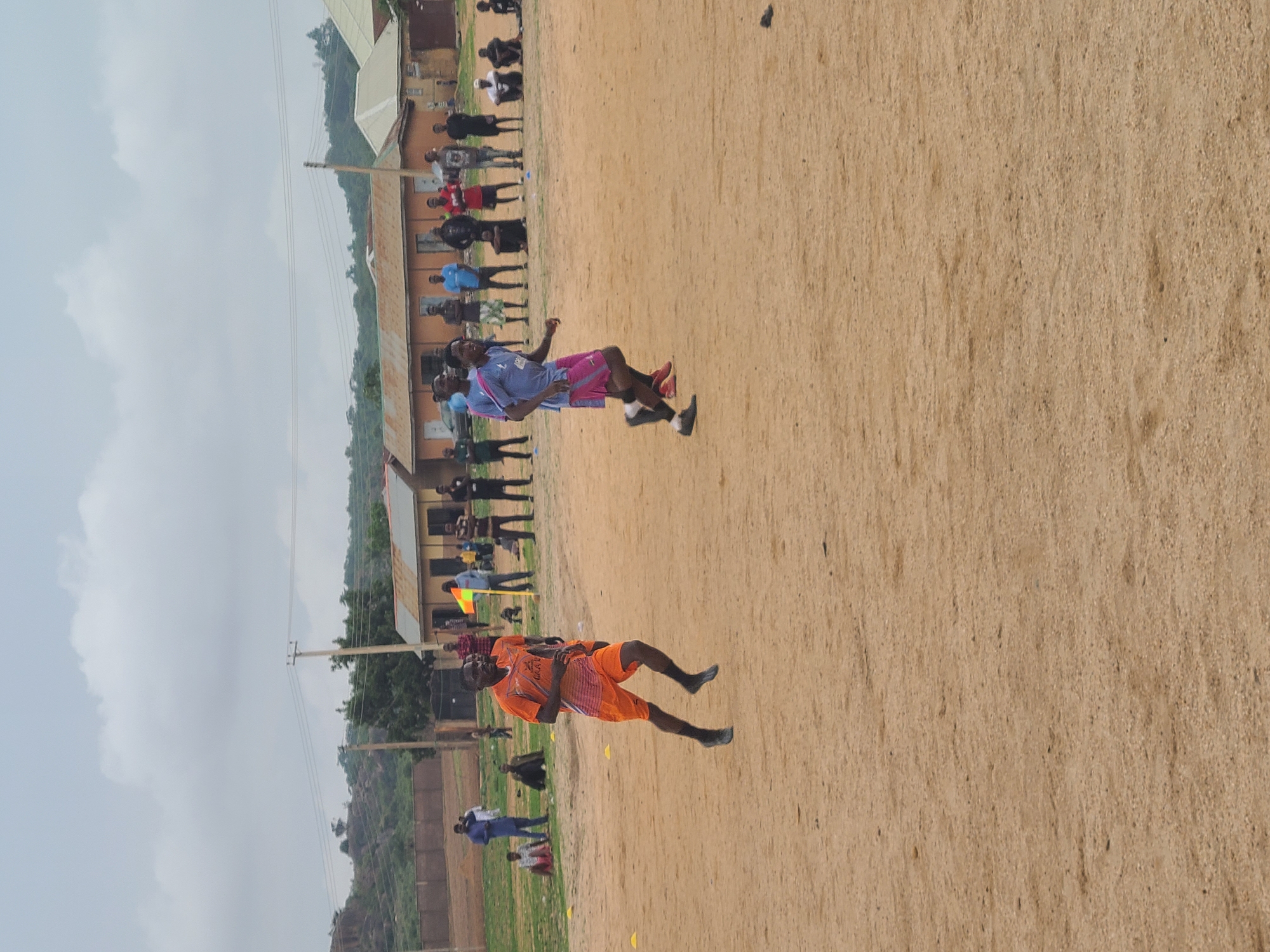 Two soccer players, one in an orange uniform and the other in a blue uniform, are on a dirt field. A crowd of people watches in the background, near a building with a corrugated roof. The location appears to be a soccer field in an unspecified city. The sky is overcast.