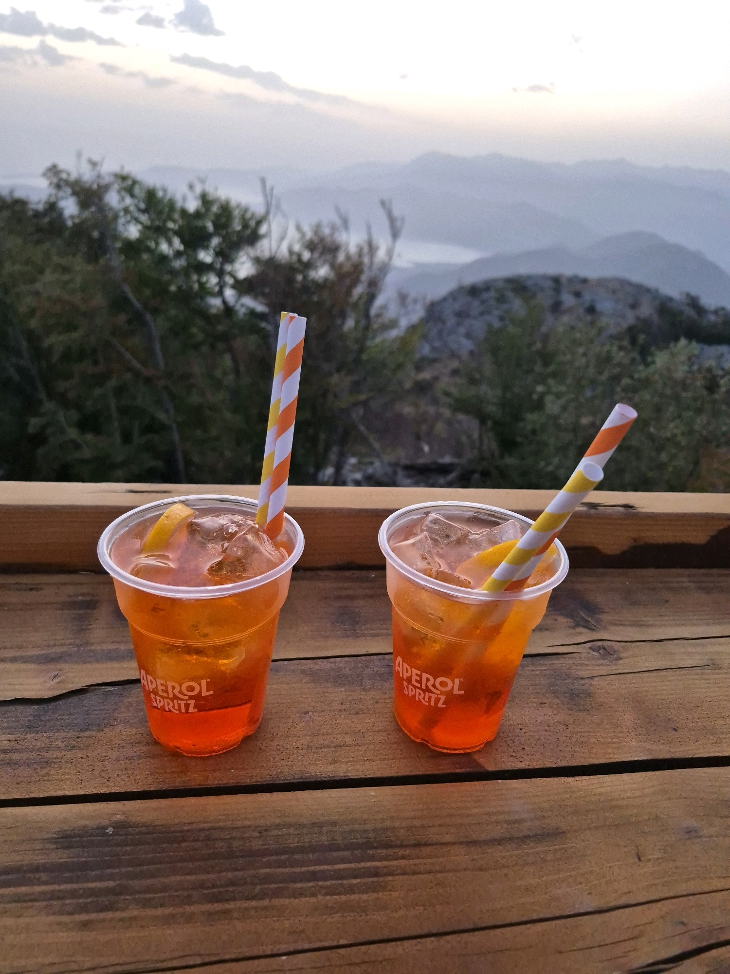 Two Aperol Spritz cocktails with orange slices and striped straws are placed on a wooden surface, likely a table or railing. In the background, a misty mountainous landscape with a lake or body of water is visible under a pale sky. The drinks appear to be in plastic cups with the "APEROL SPRITZ" logo. The scene is set outdoors, suggesting a scenic overlook or a bar with a view. The lighting indicates it might be dusk or early morning. The location is identified as Kuk, Montenegro, a mountainous region known for its natural beauty. There are no people visible in the image.
