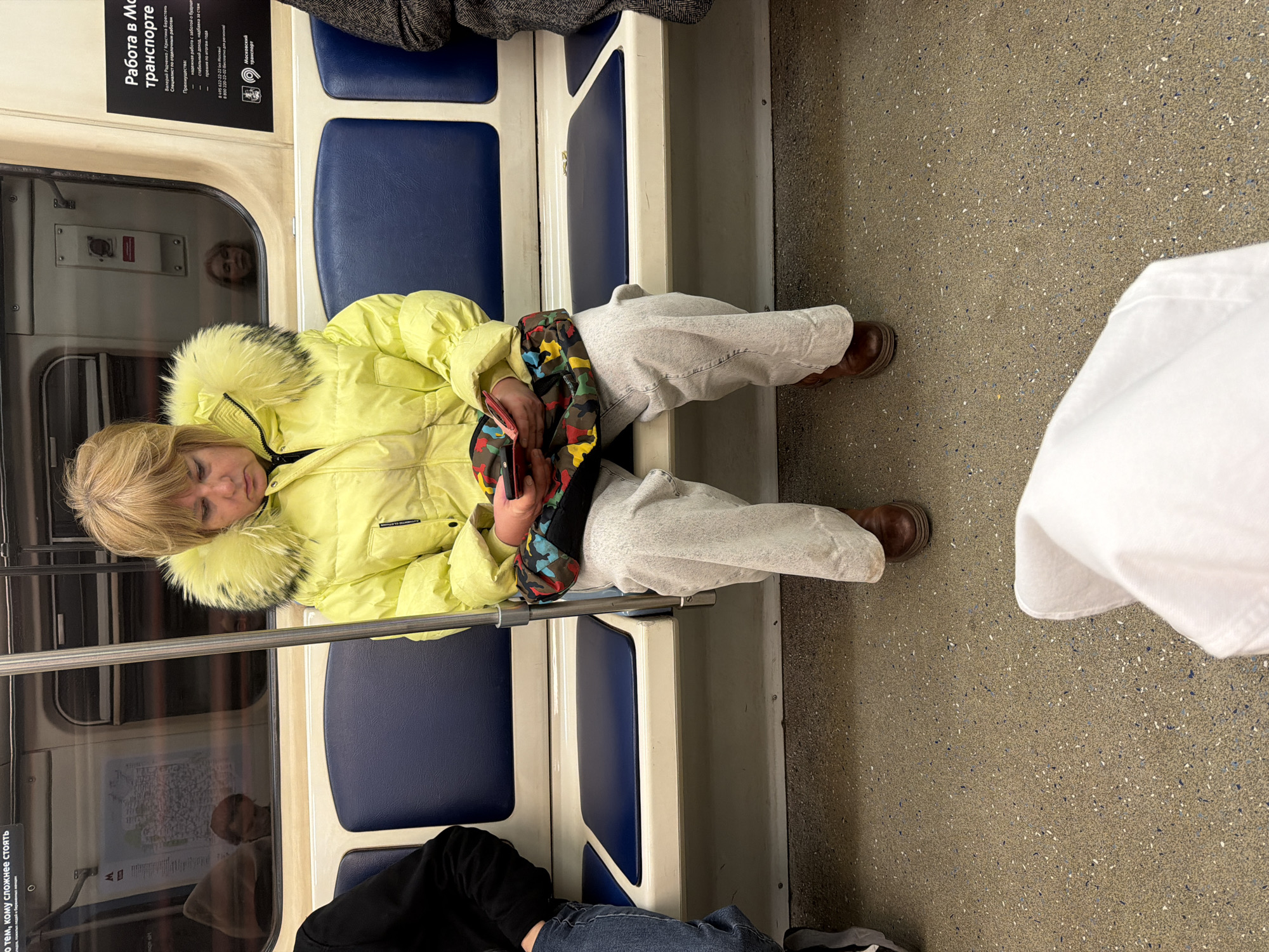 The image captures a scene inside a Moscow Metro train. The primary subject is a woman with blonde hair, wearing a bright yellow winter jacket with a prominent fur-lined hood, light-colored sweatpants, and brown shoes. She is seated on a blue upholstered bench with white frames, leaning against the train wall or window, appearing to be asleep or resting with her eyes closed. A colorful, patterned bag, possibly a fanny pack, is draped over her lap, and she holds a smartphone in her hands.
The setting is a typical metro carriage, featuring blue and white seating, a speckled gray floor, and metallic grab bars running horizontally above the seats. Reflections of other passengers are visible in the train window beside the woman, including a face looking towards the viewer and what appears to be a metro map further down the carriage. The legs of another passenger, wearing dark pants and jeans, are visible in the lower foreground.
Prominent Russian text on a black and white advertisement sign above the seats reads: "Работа в Московском транспорте" (Work in Moscow transport). Below this are names "Валерий Радченко / Кристина Берестень" and a list of "Преимущества" (Advantages) for working in transport, including "надежная работа с заботой о будущем" (reliable work with care for the future), "стабильный доход, надбавка за стаж" (stable income, seniority bonus), and "премия по итогам года" (annual bonus), along with phone numbers and the "Московский транспорт" (Moscow transport) logo. Another partially visible sign reads "но тем, кому сложнее стоять" (but for those who find it harder to stand), indicating reserved seating. The overall atmosphere is quiet, typical of a public transport commute where passengers are resting.