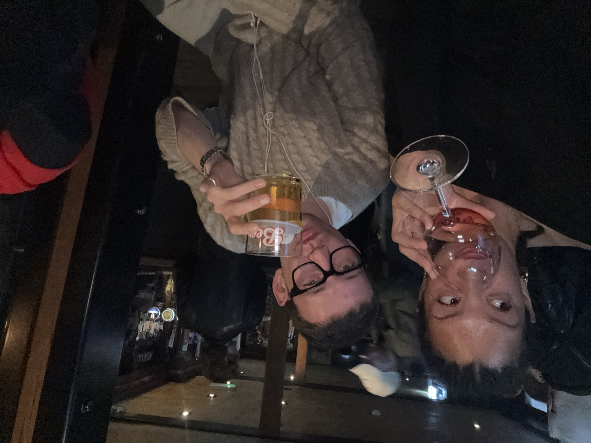 An upside-down, dim-lit indoor image features two individuals, a man and a woman, positioned in the lower half of the frame with their heads near the bottom edge, looking upwards towards the camera.
The man, on the left, wears dark-rimmed glasses and a light-colored, textured or cable-knit sweater. He holds a clear glass filled with an amber liquid, likely beer, which has the word "Beer" printed in white on its side. Wired earphones are draped around his neck and chest. He also wears a ring on his finger and a bracelet on his wrist. His eyes are directed towards the camera with a neutral expression.
The woman, on the right, holds a stemmed glass containing a pinkish-red liquid. She has dark hair and appears to be wearing a dark top or jacket, with earrings visible. Her gaze is directed slightly off-camera.
The background is dark and blurry, suggesting a dimly lit indoor setting, possibly a bar or pub, given the drinks. A dark wooden vertical beam is prominent on the left side. In the deeper background, faint glowing signs or posters are visible. Several small, illuminated spots are scattered across what appears to be the floor or a reflective surface beneath the subjects. The overall atmosphere suggests an evening social outing.