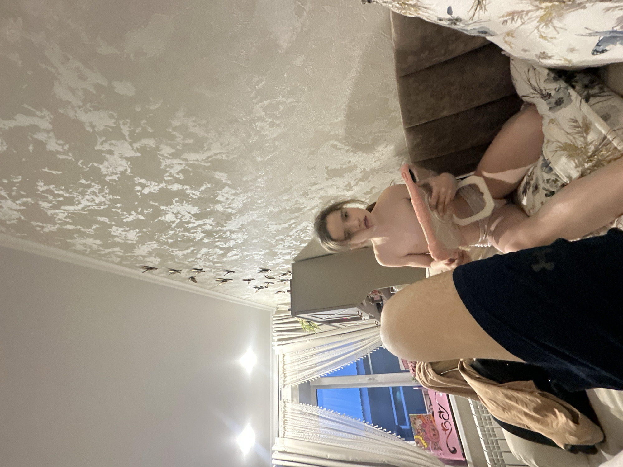 Here is a forensic description of the image:
A woman is lying on a bed holding a pink vibrator and a white object that appears to be a breast pump flange. She is partially undressed. The bed has a brown upholstered headboard and floral-patterned bedding. The room has white walls and a ceiling with a textured finish. A window with white curtains is visible in the background, letting in natural light. There are decorative bird-shaped objects attached to the ceiling near the wall. Other personal items can be seen near the window, including what appears to be a painting. The location is an interior space, likely a bedroom in an unknown city. Another person is partially visible at the bottom right of the frame.