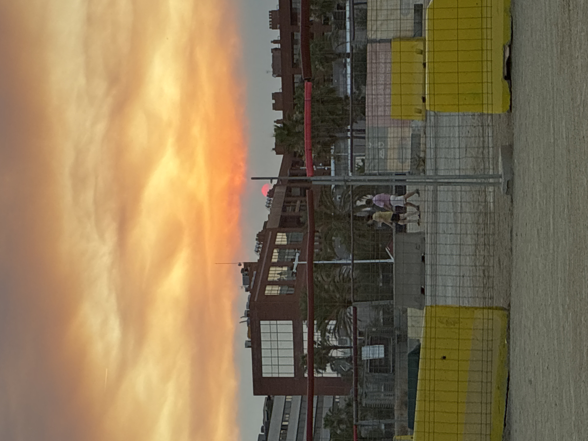 The image depicts a dusky scene in what appears to be a coastal urban area, likely Barcelona, Spain, given the architectural style and palm trees. In the foreground, there are temporary yellow barriers and wire fencing, suggesting construction or event setup. Beyond this, two individuals, possibly tourists or locals, are walking. In the middle ground, several modern buildings with glass facades are visible, interspersed with palm trees. A distinctive reddish building with large, multi-paned windows stands out. The sky is dominated by a vibrant sunset, with hues of orange, yellow, and red, and the sun itself is a small, bright red orb partially obscured by the horizon. A faint contrail from an aircraft is visible in the upper left portion of the sky. The overall impression is of an urban environment at the end of the day, with a dramatic sky overhead.