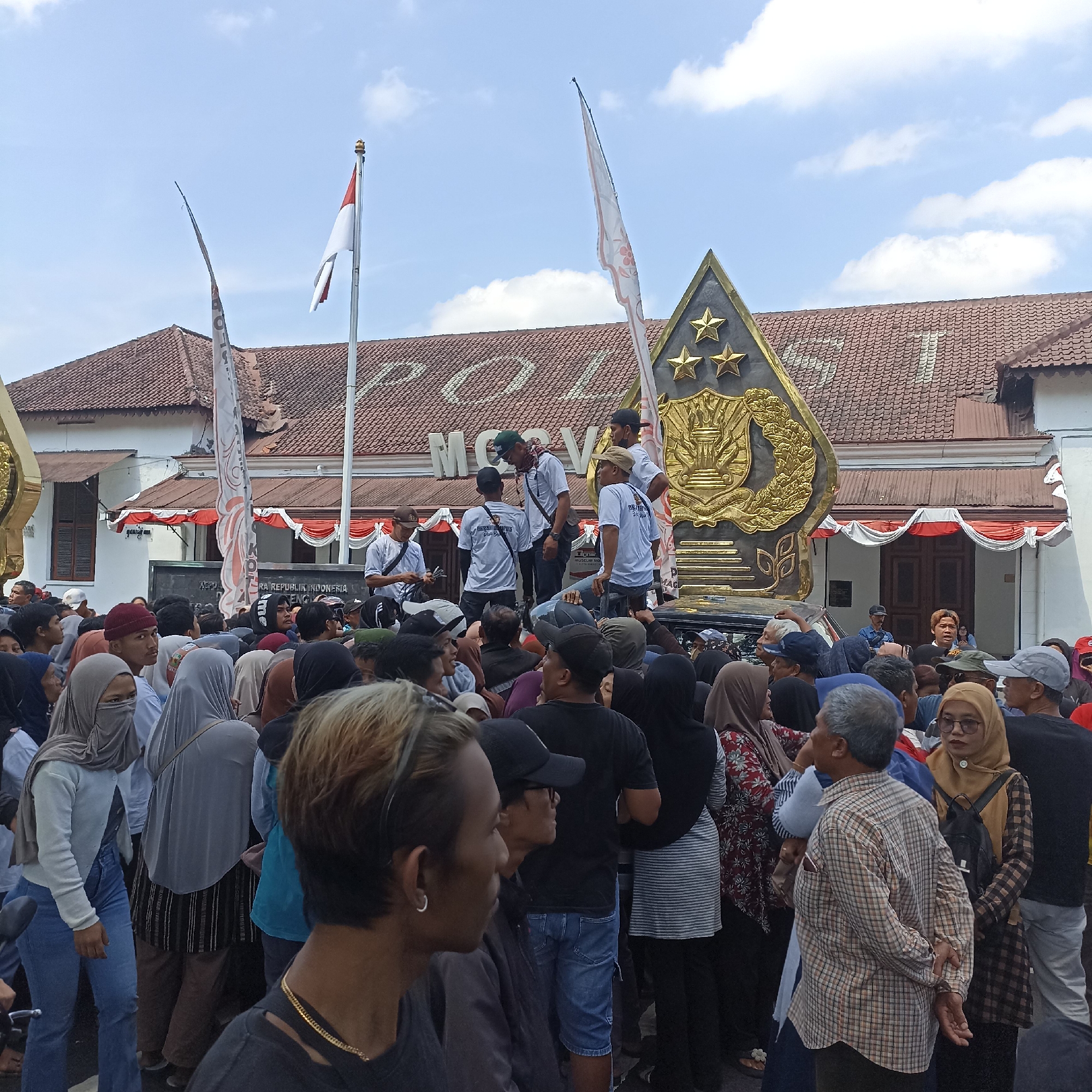 In the city of Magelang, Indonesia, a crowd of people has gathered in front of a building with a tiled roof. The building has "POLRI" visible on its facade, indicating it is a police station. Two flags are flying, one is red and white, and another white flag with red intricate patterns. A large golden crest with three stars is prominently displayed in front of the building. Many people are dressed casually, with some wearing t-shirts and jeans, while others are wearing headscarves. The sky is blue with scattered clouds. The atmosphere appears to be that of a public gathering or event.