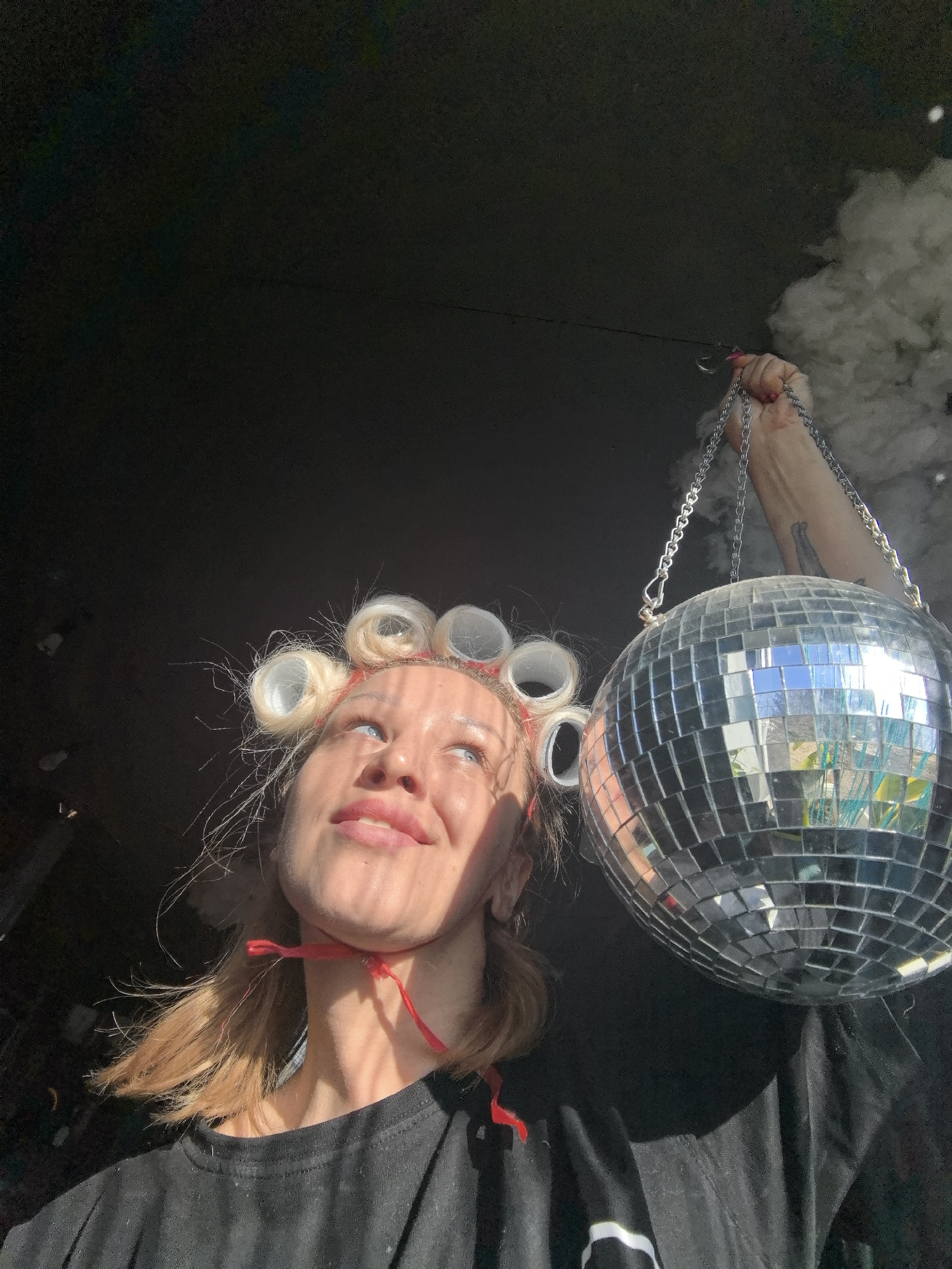 A woman stands indoors, looking upwards with a slight smile. She has blonde hair with curlers, and a red ribbon around her neck. She is holding a disco ball by its chain with her right hand. The background is dark, with a patch of smoke or haze visible on the right side. It is not possible to ascertain the location of this scene without further information.