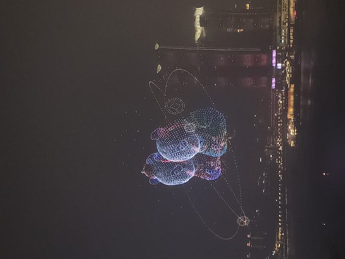 The scene, captured at night, displays an aerial drone light show above a body of water, set against the illuminated skyline of Jiangbeicheng Subdistrict, Chongqing, China. Two large, volumetric figures resembling pandas are formed by numerous small, multicolored lights (white, blue, pink, green) in the dark sky, appearing to embrace. A luminous, dotted elliptical pattern encircles the figures, with smaller light clusters along its path. Faint reflections of the display are visible on the dark water below. The far bank is characterized by a dense array of brightly lit high-rise buildings and skyscrapers, including one with a distinctive wavy illuminated roofline. Several illuminated vessels are present on the water near the city's edge. No individuals are discernible within the frame.