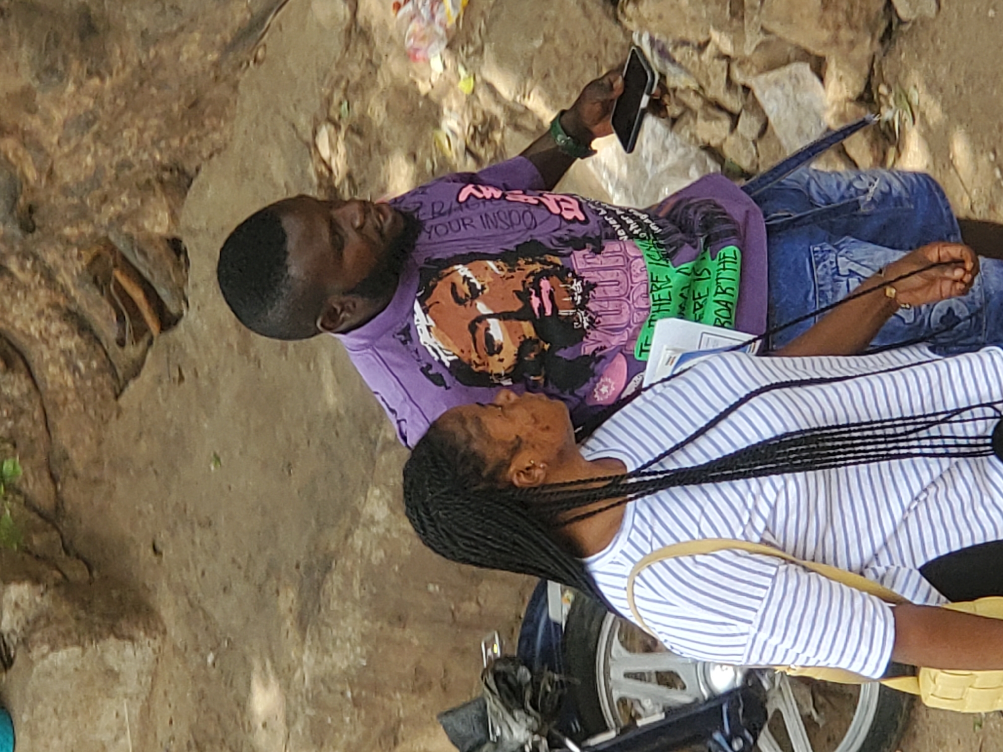 In Jos, Nigeria, a man and a woman are positioned on or next to a motorcycle. The man is lying down and looking at his phone, he is wearing a purple graphic T-shirt and blue jeans. The woman is lying down with her head near his torso and she is wearing a striped shirt. They are outdoors on a concrete surface.