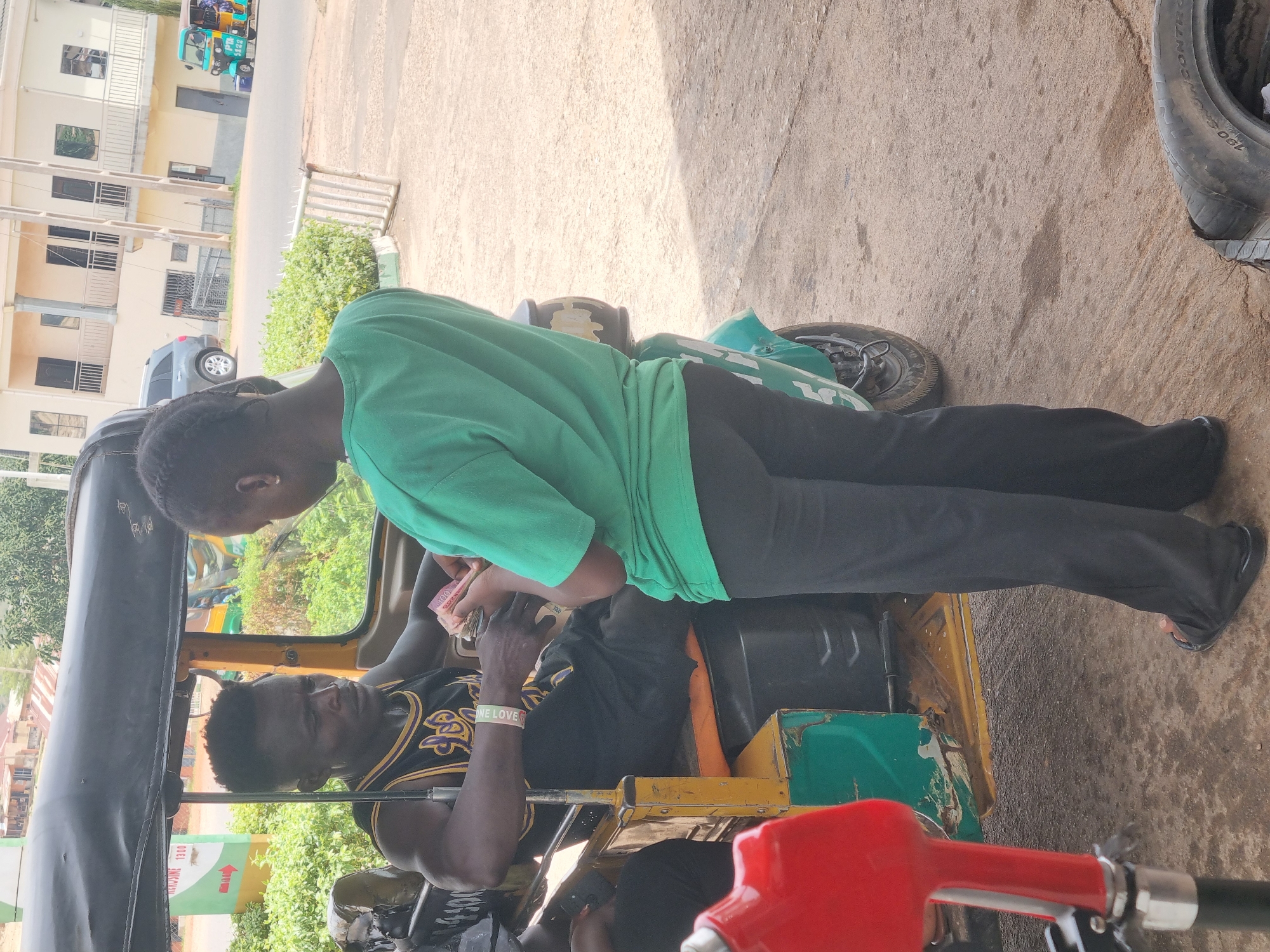 The image captures a scene in Jos, Nigeria, focusing on an interaction at what appears to be a gas station. A person in a green shirt and black pants is seen handing money to a man seated inside a yellow and green tricycle. The man inside wears a black and yellow jersey and a wristband with the words "ONE LOVE". The tricycle is parked near a gas pump with a red nozzle. In the background, there are buildings and greenery, typical of an urban environment. A tire is visible on the pavement, hinting at a repair or tire service area.