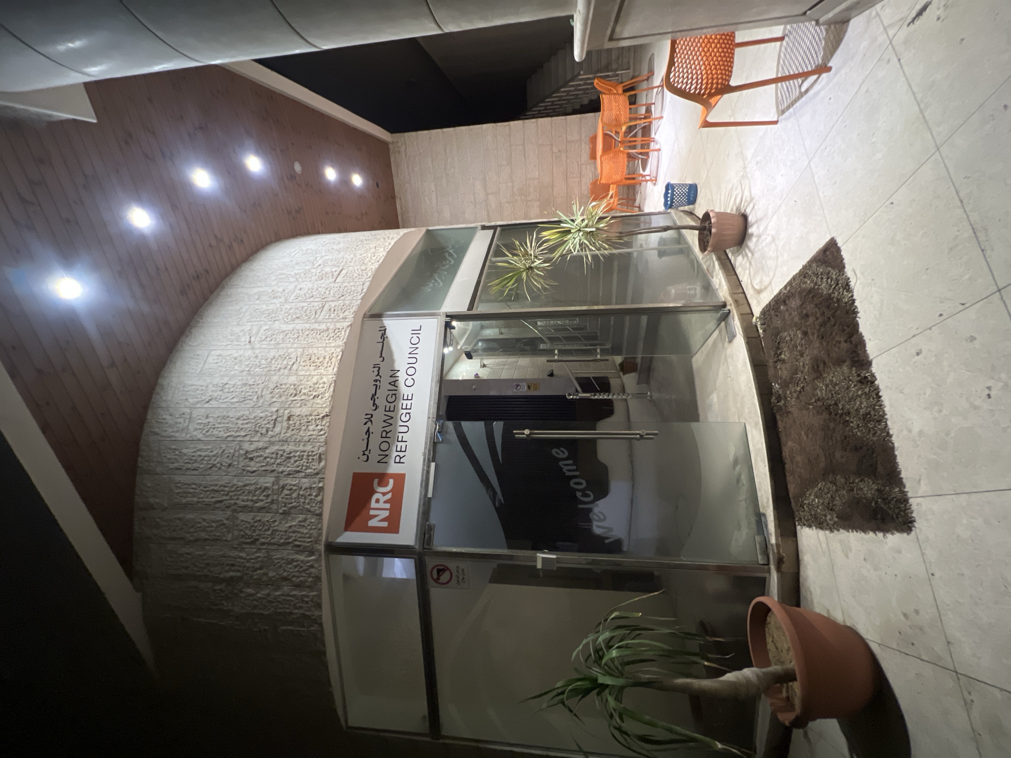 The image shows the entrance to the Norwegian Refugee Council (NRC) office in Gaza City, Palestinian Territories. The NRC logo is prominently displayed on the glass facade, alongside Arabic text. The entrance area features a glass door with metal handles, and a "welcome" inscription on the interior glass. Outside the entrance, there is a potted plant on the left and a dark mat on the floor. To the right of the entrance, there is an outdoor seating area with orange chairs and tables. Further to the right, a staircase leads upwards, partially visible. The building has a curved, textured facade and a wooden-paneled ceiling with recessed lighting. The overall scene is lit by artificial lights, suggesting it is nighttime or a dimly lit indoor space.