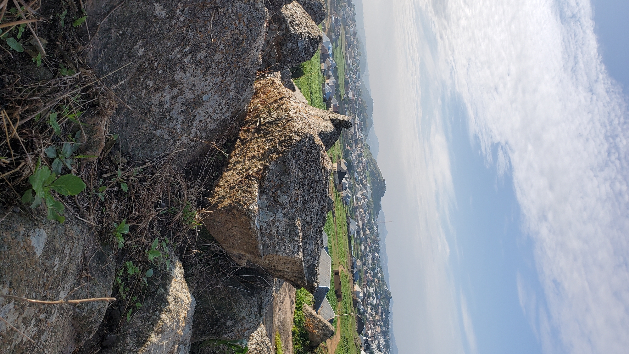 This image captures a serene, wide-angle view of a settlement nestled amongst rolling hills under a partly cloudy sky. In the foreground, large, weathered boulders dominate the scene, their rough textures and varied shades of gray hinting at their age and the geological landscape of Jos, Nigeria. Dry grass and small green shoots sprout from the rocky terrain, adding a touch of life to the rugged foreground.
Beyond the rocks, the landscape unfolds into a patchwork of urban and rural elements. Rows of houses, characterized by their diverse roof colors and architectural styles, spread across the hillsides. Many homes have metallic roofs, reflecting the sunlight. Interspersed between the houses are patches of vibrant green farmland, suggesting agricultural activity within the settlement. The overall impression is one of a thriving community integrated with its natural environment. The distant hills, shrouded in a light haze, provide a picturesque backdrop, emphasizing the expansive nature of the vista. The light suggests it is either early morning or late afternoon, with the sun casting soft shadows.