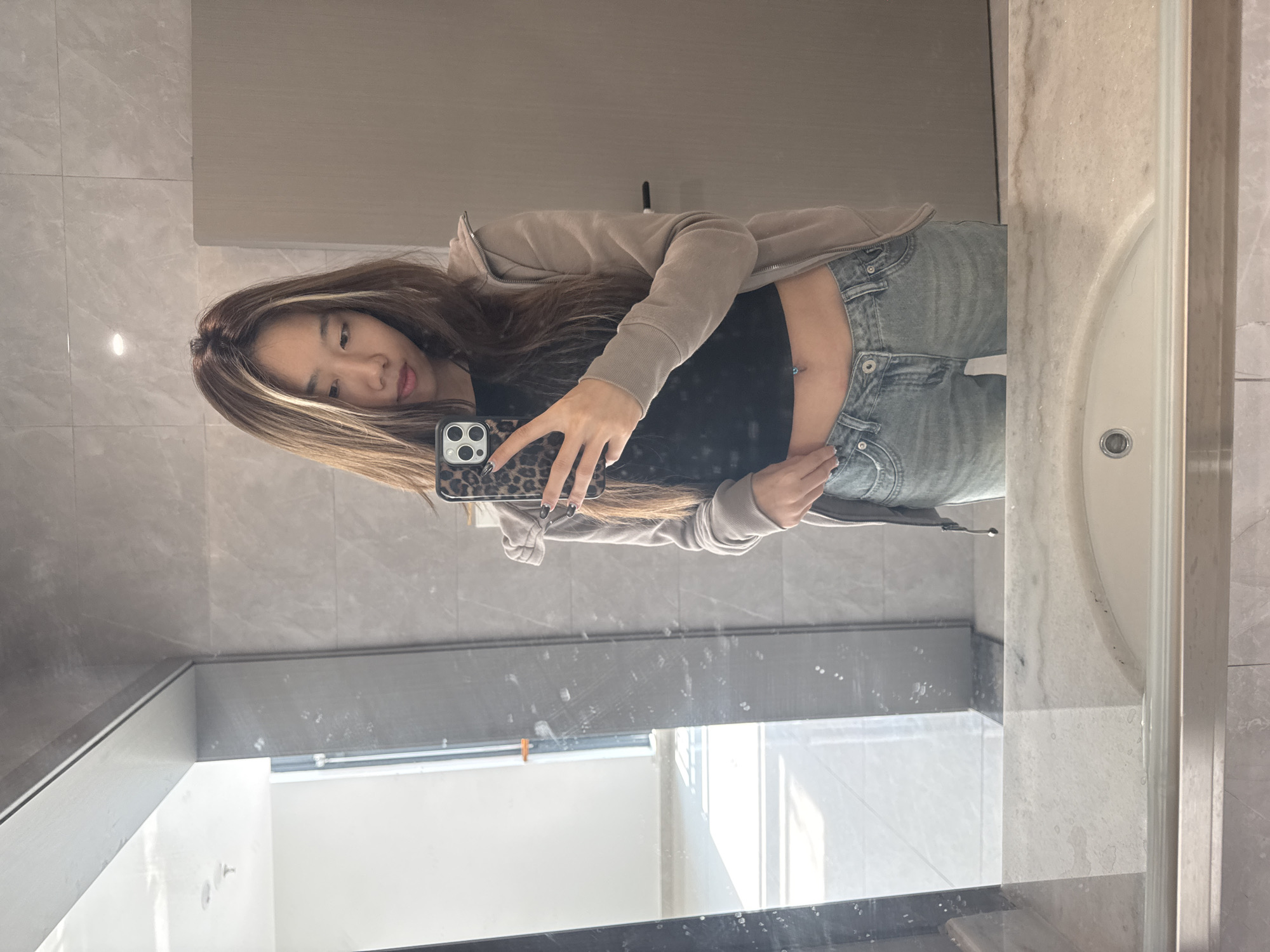 A young woman stands in a bathroom in Nan'an District, China, taking a mirror selfie. She has long brown hair with blonde highlights and is wearing a black top, light-wash jeans, and a beige zip-up hoodie. Her phone has a leopard-print case, and her nails are painted black. The bathroom features grey tiled walls, a white sink, and a window.
