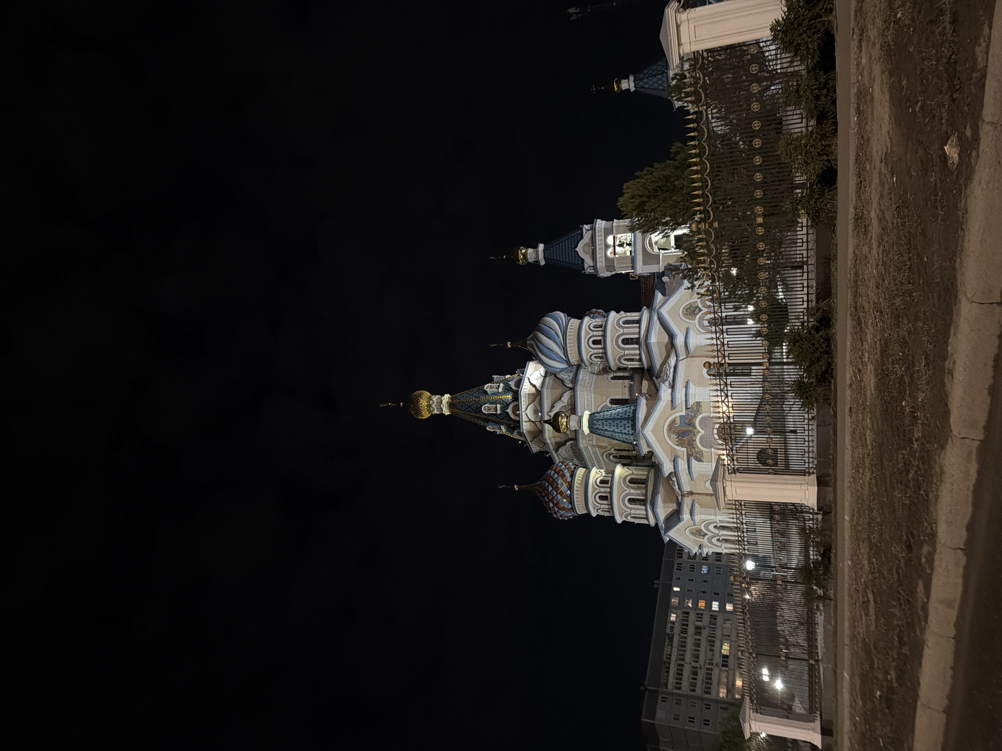 The image shows the Cathedral of St. Michael the Archangel in Izhevsk, Russia at night. The cathedral, with its distinctive domes and ornate architecture, is illuminated against the dark sky. It is situated behind a decorative metal fence, and a building with lit windows is visible nearby. A paved area and some greenery are also present in the foreground.