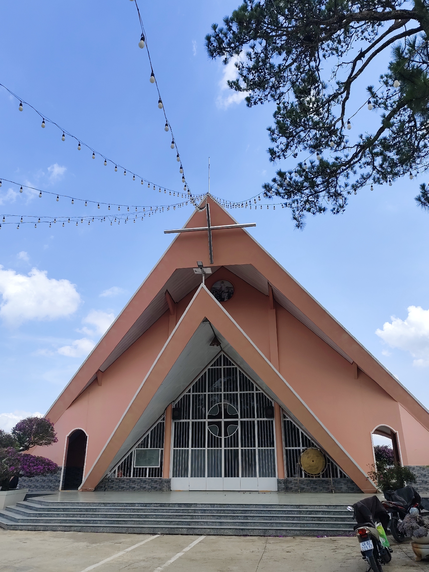 The image depicts the Chicken Church (Nhà thờ Con Gà), a landmark located in Đa Thọ, Da Lat, Vietnam. The pink church has a triangular facade, with a central entrance featuring white doors and a glass design. A cross is positioned at the peak of the roof. Strings of lights are suspended in the air. To the left of the church, a flowering shrub is visible. To the right, a parked motorcycle is partly obscuring a flower pot. The sky is light blue with scattered clouds. Steps lead up to the church entrance.