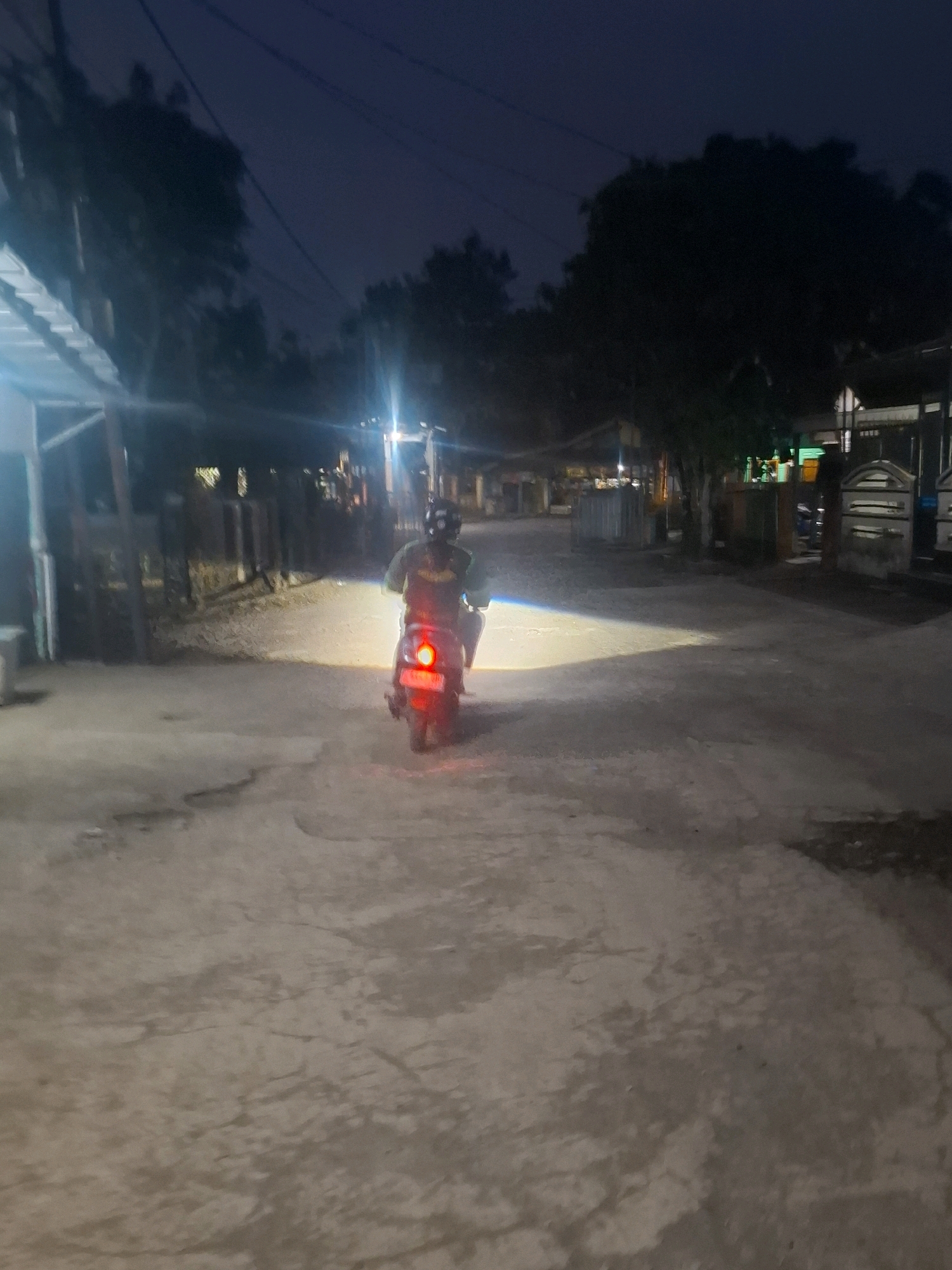 A lone individual on a scooter rides down a paved road at dusk in Ciheulang Tonggoh, Indonesia. The scooter's headlight illuminates the path ahead, casting a bright beam across the rough, uneven road surface. The rider is wearing a helmet and a reflective vest, suggesting they are either a local resident or a delivery person. The surrounding environment appears to be a residential area, with low-lying buildings and trees visible in the background. Streetlights can be seen in the distance, adding to the dimly lit atmosphere. The sky is a deep blue, indicating the transition from day to night. The overall impression is one of a quiet, peaceful evening in a rural Indonesian village.