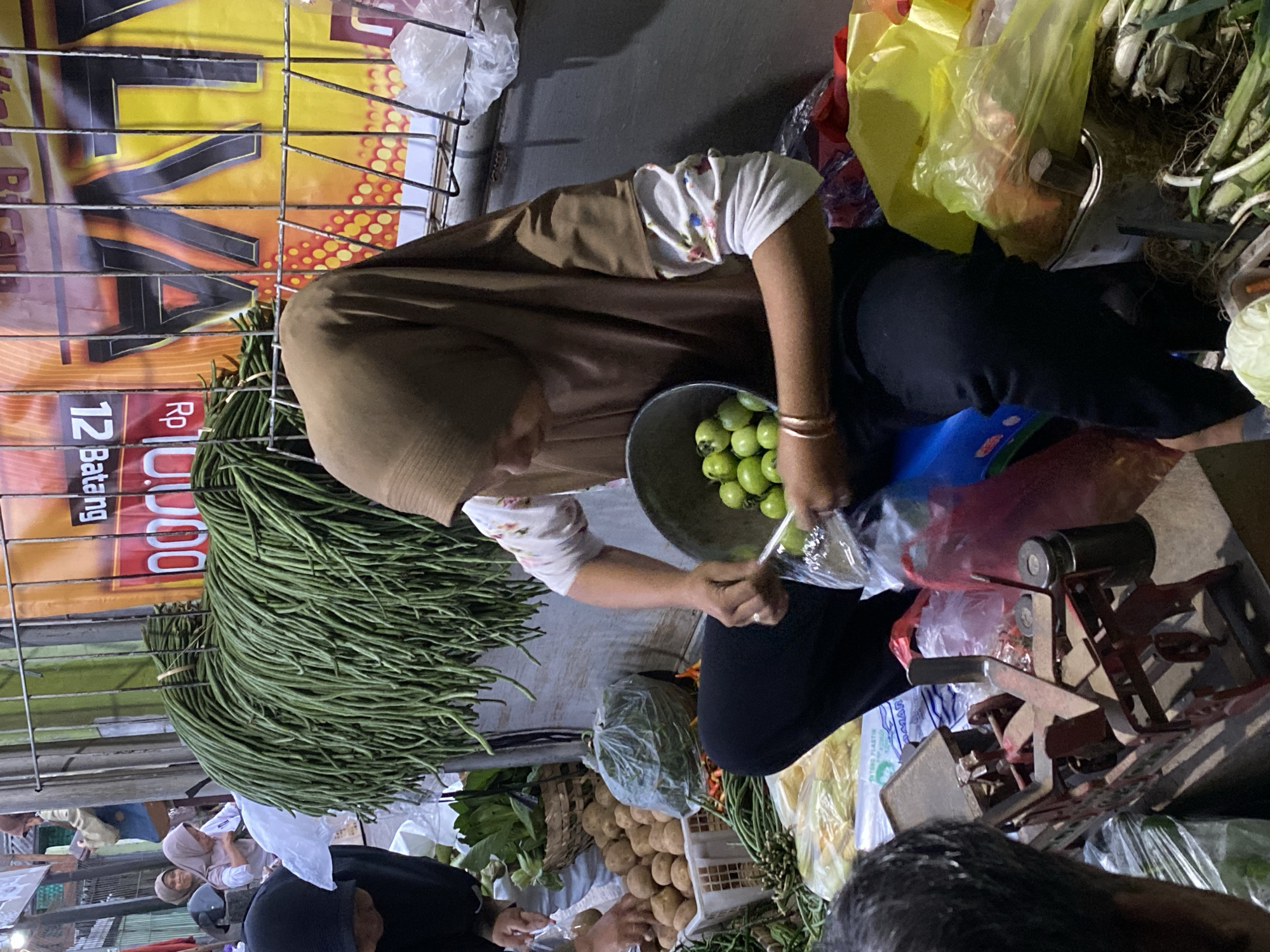 This is a medium shot, captured at a street market stall in Ambarketawang, Indonesia. The scene depicts a woman wearing a brown hijab and a floral-patterned long-sleeved shirt, along with black pants. She is seated and actively sorting small, green, round fruits from a metal bowl into a clear plastic bag, using a utensil. In the foreground, a red and metal weighing scale is visible, and to the left, another person's shoulder and head are partially in frame, with their back turned to the camera.
Behind the woman, a large bundle of long, green beans hangs prominently, supported by a metal grate. Other produce, including potatoes in a white crate and a bag of leafy green vegetables, are also visible. In the background, a large banner with Indonesian text and graphics adds context to the market setting. The overall lighting suggests an outdoor environment, possibly at dusk or in a shaded area.