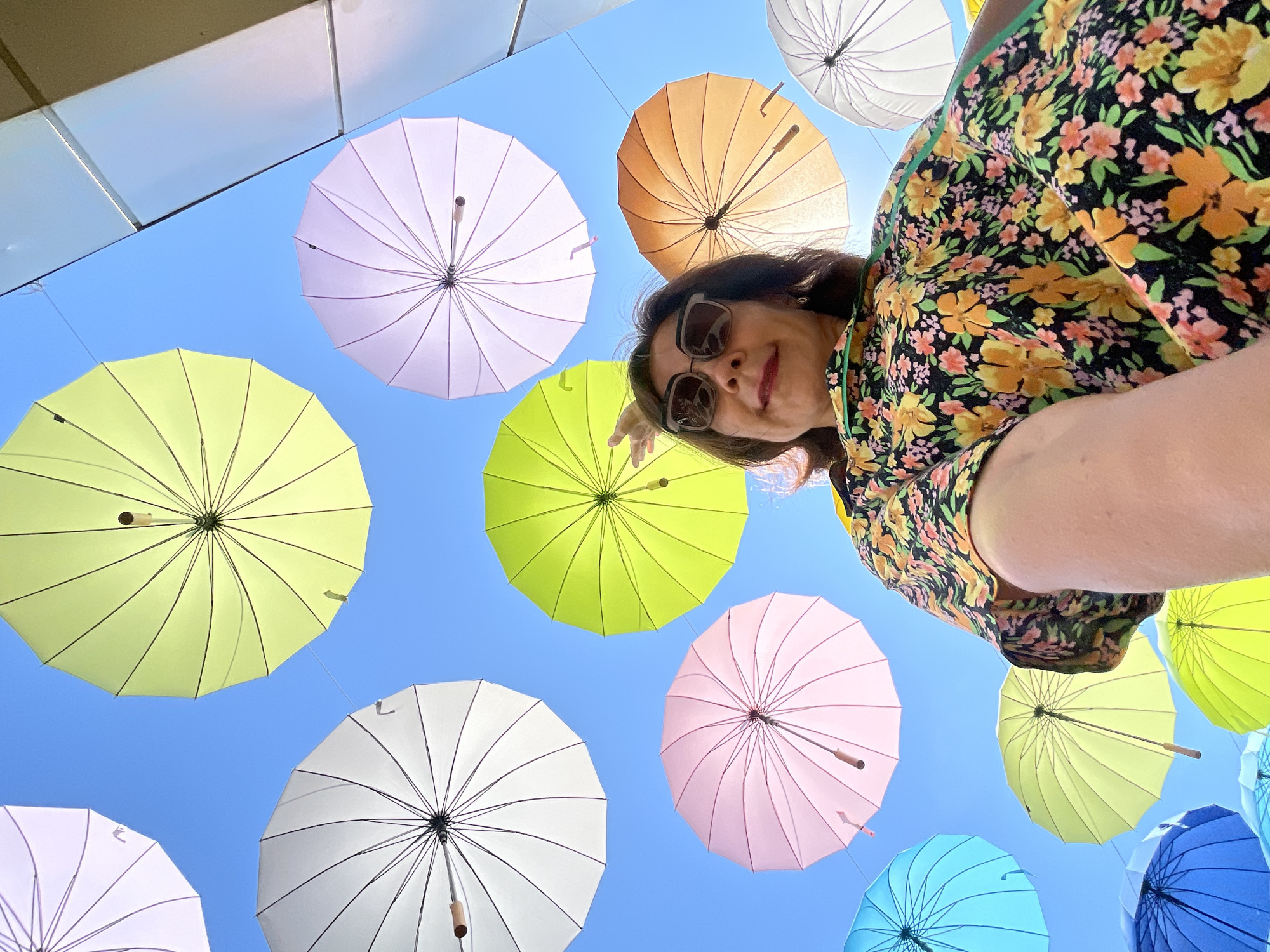 An over-the-shoulder, low-angle shot captures a woman posing in front of a vibrant display of colorful umbrellas suspended against a bright blue sky. The woman, with dark hair and sunglasses, wears a floral-patterned blouse. The umbrellas, in various hues of pink, yellow, white, orange, and blue, are spread out above, creating a visually striking canopy. This scene is located in the city of Sochi, Russia, a popular resort destination known for its picturesque landscapes and recreational activities.