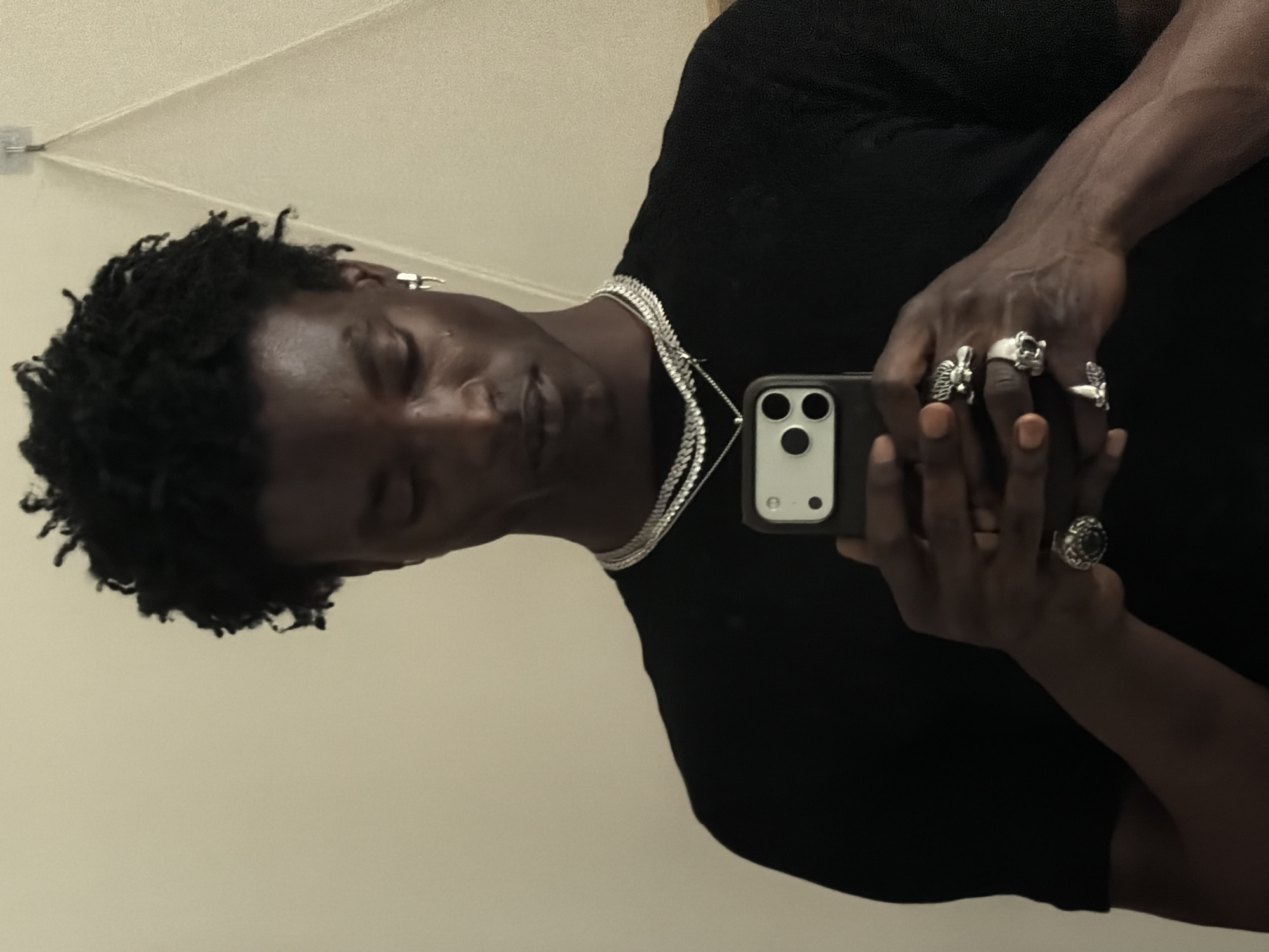 A man with dark skin and short, dark curly hair is holding a smartphone to take a selfie. He is wearing a black t-shirt and two silver necklaces. He also has multiple silver rings on his fingers. The background is a plain, light-colored wall. There is no indication of the location being London, United Kingdom.