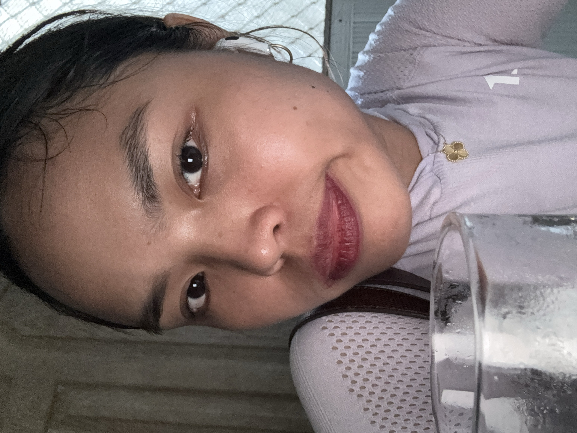 A close-up, selfie-style portrait captures a woman's face from the chest up, with a glass of water prominently in the foreground. The woman has dark hair, styled away from her face, and her eyes are open and looking towards the camera. She is wearing a light purple, long-sleeved top with a mesh-like texture on the shoulders and a small, gold clover-shaped pendant. Her lips are slightly parted, and she appears to be holding the glass of water. The background is indistinct, suggesting an indoor setting, possibly a home or hotel room. The lighting is somewhat dim, with natural light filtering in from a window on the right side of the frame. The overall impression is casual and personal.
The image does not contain any visual cues that would allow for specific identification of the location within Davao City, Philippines.