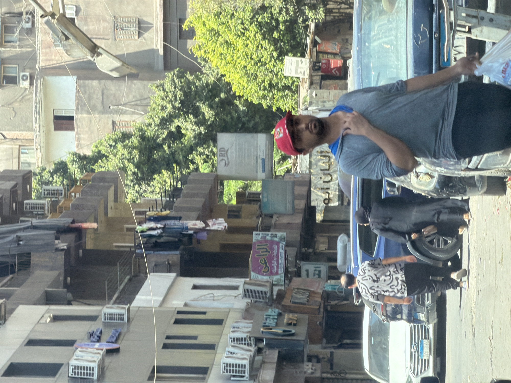 The image depicts a street scene in Cairo, Egypt, characterized by multi-story buildings lining a street. The architecture features balconies, air conditioning units affixed to walls, and laundry hanging from lines. Several businesses with signs in Arabic are visible at street level.
In the foreground, a man wearing a red baseball cap and a gray hooded shirt is walking towards the viewer, carrying a clear plastic bag. Behind him, two individuals are walking away from the camera, one in a patterned shirt and dark pants, the other in a black abaya. Several vehicles, including cars and what appears to be a public transport bus, are parked or moving on the street. The overall impression is of a busy urban environment with a mix of residential and commercial activity.