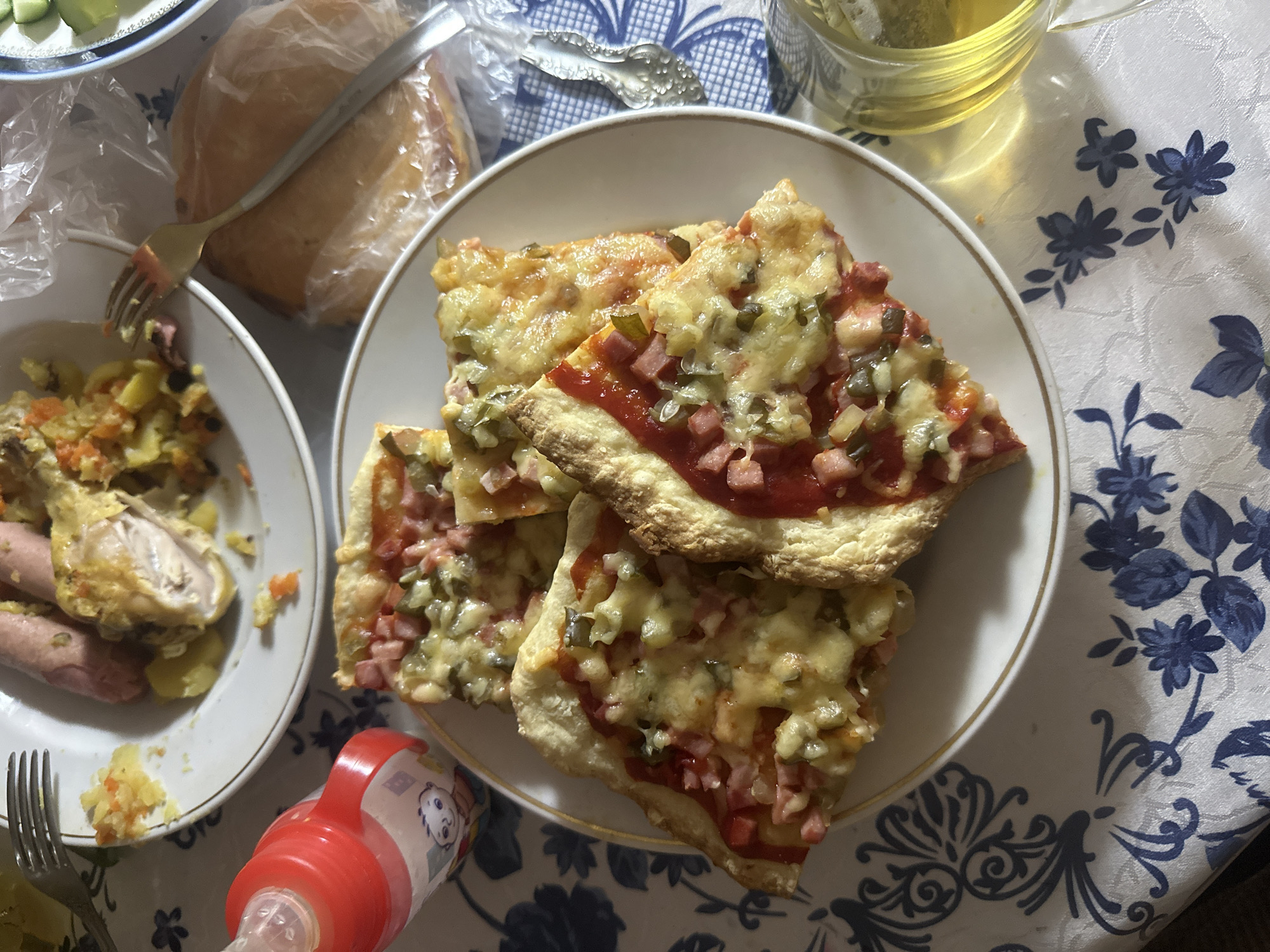 On a tablecloth with a blue floral pattern, there is a plate of square pizza slices topped with tomato sauce, diced ham, pickles, and cheese. Next to it, there's a plate containing a chicken leg, sausages, and some cooked vegetables. A baby bottle with a red lid sits near the pizza. Bread in plastic wrap is also present, along with a clear glass mug filled with a light-colored beverage. The setting appears to be a home environment in the city of Unknown, Unknown.