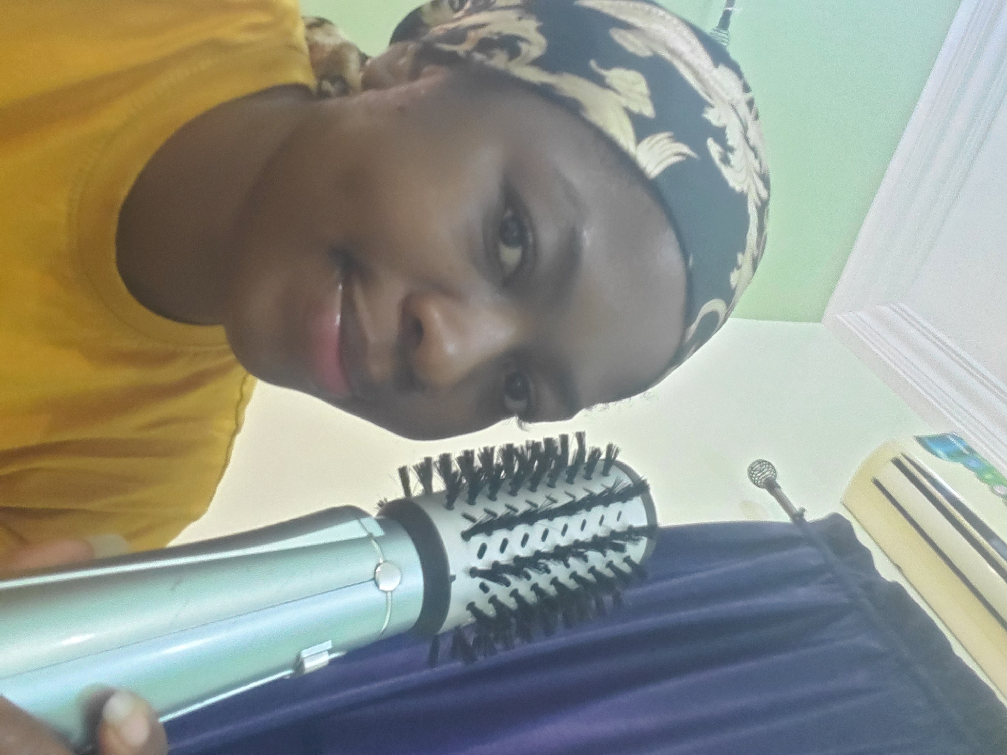 A dark-skinned individual, appearing to be female, is shown in a medium shot, facing the viewer with a slight smile. She wears a dark patterned headscarf featuring gold leaf or floral motifs and a plain yellow short-sleeved t-shirt. Her right hand holds a silver and light grey cylindrical hot air brush with a round head and black bristles, positioned to the right of her face. The background depicts an indoor setting with a light green wall on the left and a white wall on the right. A white ceiling with molded trim is visible at the top. An air conditioning unit is mounted on the upper right white wall, positioned above a dark blue curtain. A silver curtain rod with a decorative finial is partially visible against the white wall and extending into the curtain. The specific geographic location, Barden Yana, Nigeria, cannot be determined from the visual information presented.