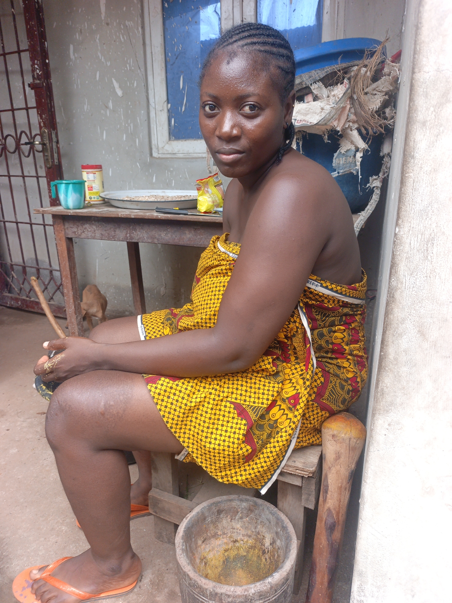 A medium shot shows a young woman seated on a wooden stool outdoors. She is wearing a yellow and red patterned wrap dress. Her hair is braided and pulled back. She has her hands clasped in her lap and appears to be looking at the camera. To her left, a wooden table holds a plate of food, a mug, and a package of snacks. Behind the table, a small, light brown dog is visible. To the right of the woman is a wooden mortar and pestle. The scene appears to be in a rural or semi-urban setting.