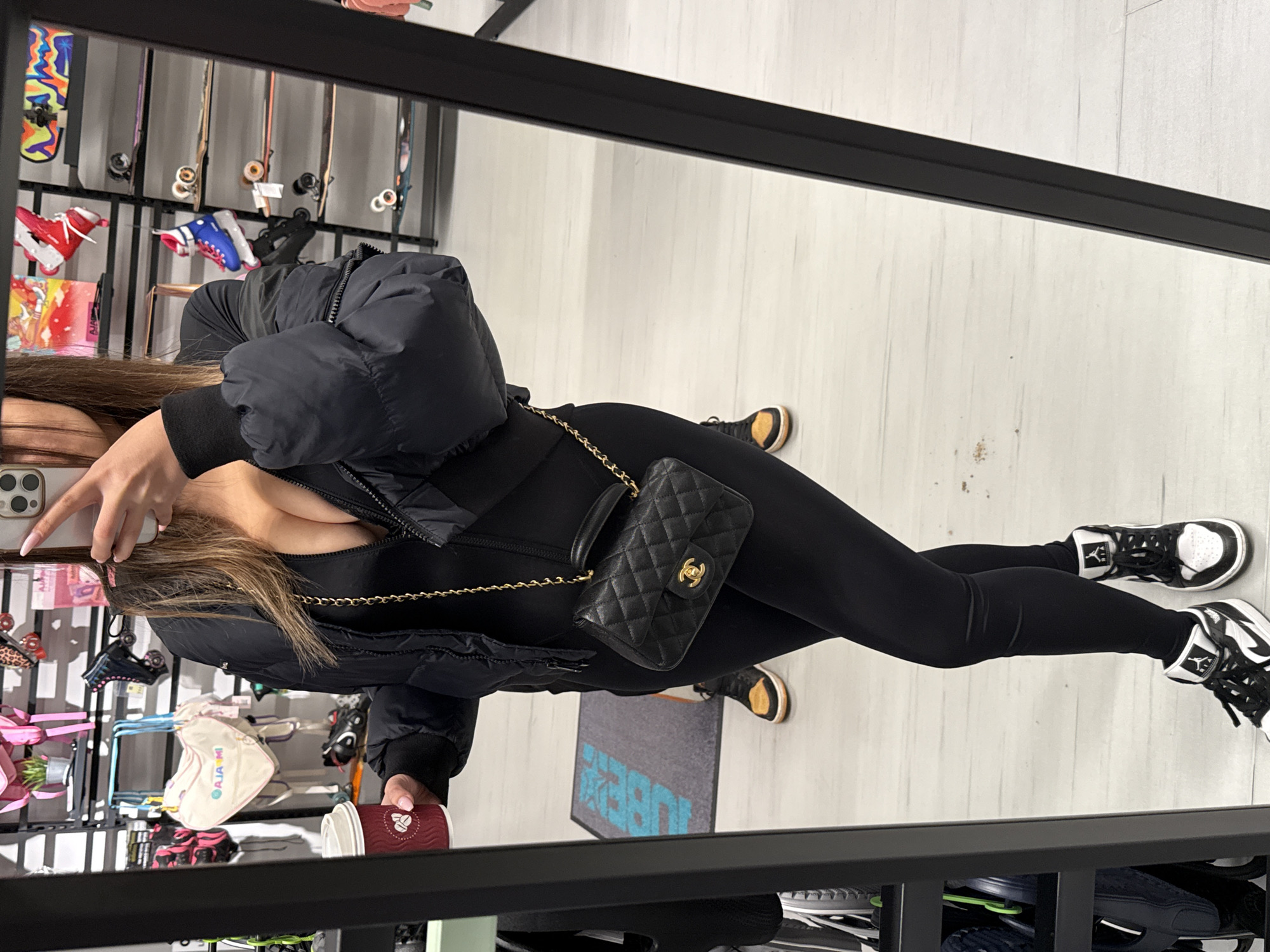 A young woman stands in front of a large mirror, presumably in a retail store. She is taking a selfie with her cell phone, holding a coffee cup in her other hand. She is wearing a black jumpsuit, a black puffer jacket, and black and white sneakers. A black quilted Chanel handbag hangs across her body. The background shows racks of skates and shoes, indicating the store's merchandise. The mirror reflects the interior of the store, including the tiled floor and various racks. It is difficult to identify the exact location in Sofia, Bulgaria, from the image alone. More details on the store's name or local landmarks would be needed to pinpoint the specific location.