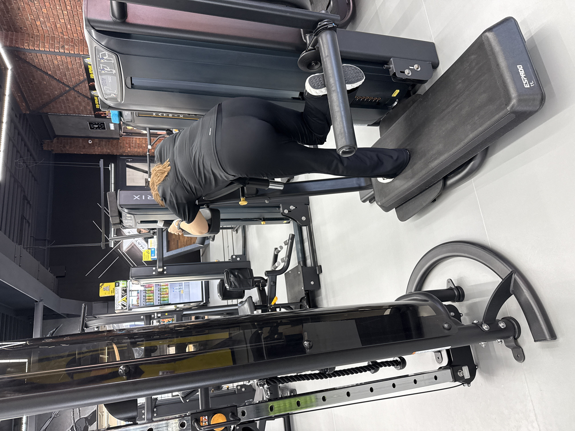 An individual is exercising at a gym in Votkinsk, Russia, using a prone leg press or glute drive machine. The person, viewed from behind, is wearing black athletic wear and has reddish-blonde hair. They are positioned face down on the machine, with their feet pushing against a large, dark grey platform labeled "ESPAADO TL" in white text. Their body is bent at the waist, indicative of a lower body workout.
The gym features modern equipment and an industrial aesthetic. Behind the person, a "MATRIX" brand treadmill is partially visible. Further back, another large fitness machine, possibly a cable station, with black and yellow accents, stands on a tiled floor. The ceiling or upper wall area on the left reveals exposed brickwork, contrasting with bright fluorescent strip lighting. In the background, a refrigerated display case for drinks is visible, with a sign above it bearing the Russian text "ФИТНЕС-БАР" (FITNESS-BAR). The overall environment is brightly lit and appears clean and well-maintained. The image is originally oriented sideways.