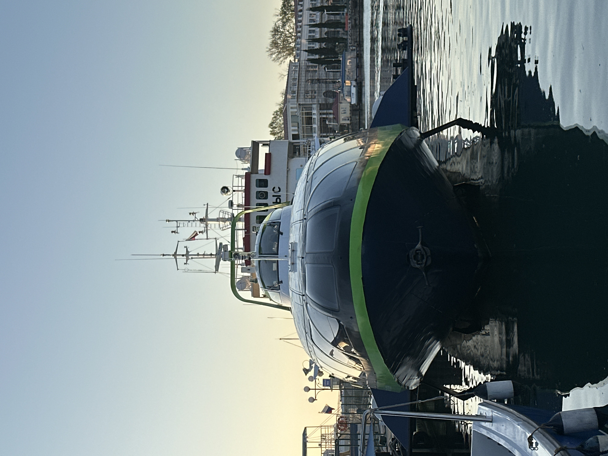 The image captures a close-up of a boat docked at a pier in Sochi, Russia. The boat is primarily dark with a green stripe running along its side. The harbor's water reflects the boat and the nearby infrastructure, a mix of other boats and buildings along the coastline. The sky suggests either early morning or late afternoon, indicated by the soft light filtering through the scene. A second boat is visible behind, with part of the Russian lettering "ЫС" is visible on the hull. In the background, buildings stand along the shore.