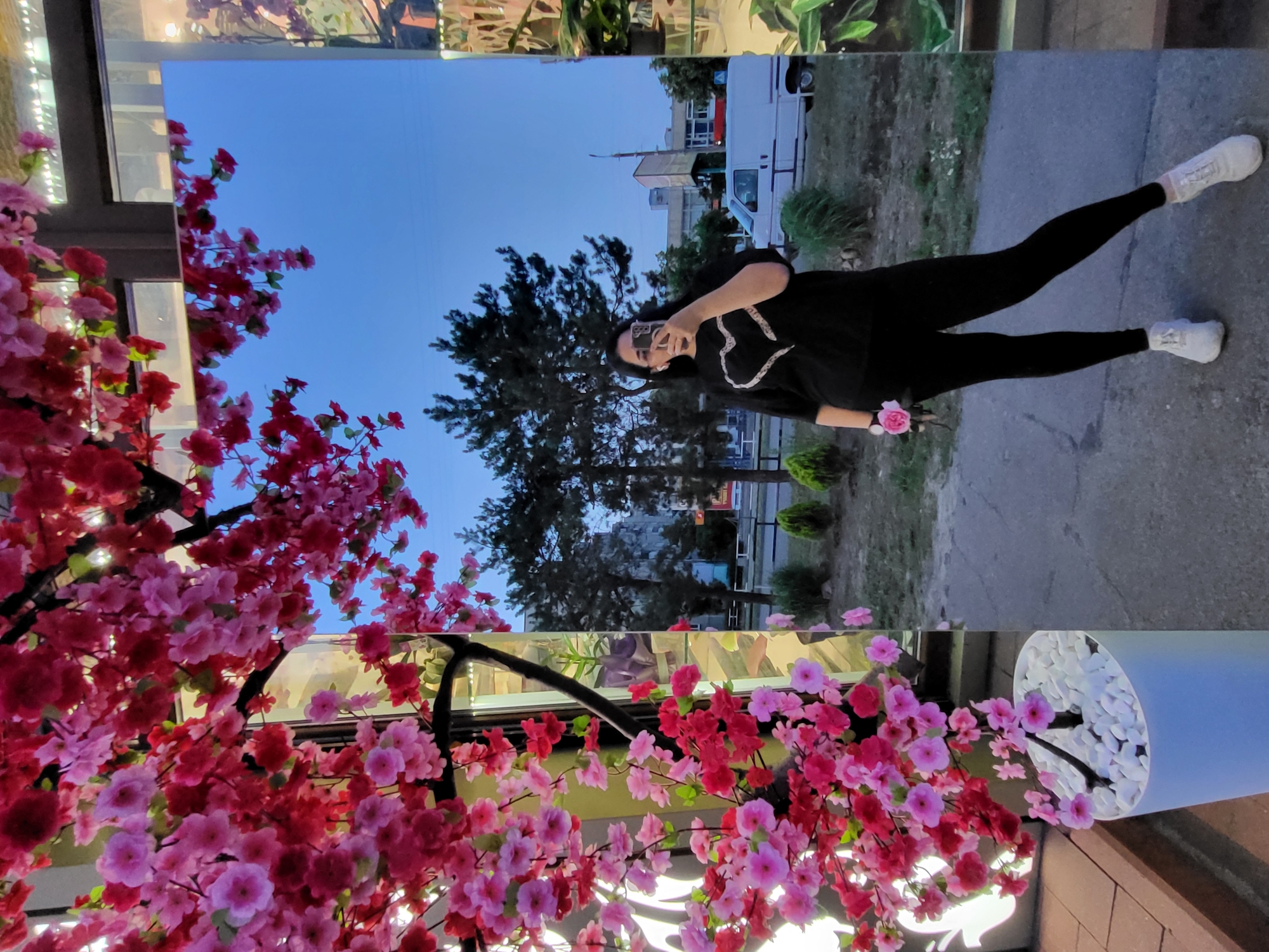A person stands in front of a mirror, taking a selfie. The person is wearing black leggings, white sneakers, and a black t-shirt with a white heart design. They are holding a pink flower in their left hand. The mirror reflects trees and buildings in the background, including a white van. The scene appears to be an outdoor setting, possibly in the city of Horishni Plavni, Ukraine, with a decorative tree with pink and red flowers in the foreground.