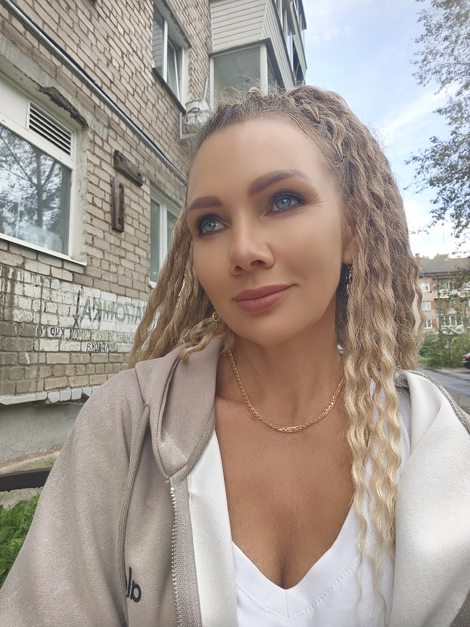 An outdoor, medium shot captures a blonde woman with blue eyes from the chest up, looking to her left, in front of a brick building. The woman's hair is styled in blonde dreadlocks, and she is wearing a tan zip-up hoodie over a white V-neck top. She is also adorned with a gold chain necklace and gold earrings. The brick building behind her is weathered, with some graffiti visible on its surface. The background also shows a tree and a cloudy sky, suggesting an urban environment.