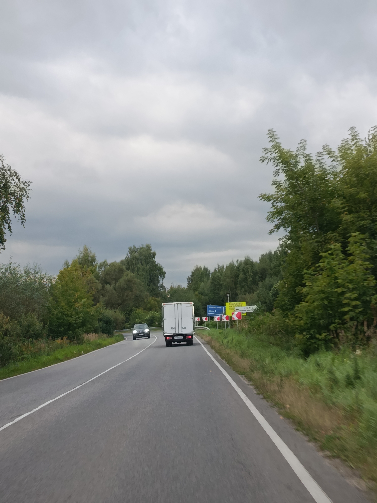 The image depicts a two-lane asphalt road with a white dividing line, curving to the right. Overcast skies dominate the upper portion of the frame. On the left side of the road, a dark-colored car is visible, its headlights illuminated. Further ahead on the right side of the road, a white box truck is traveling in the same direction. The road is flanked by lush green vegetation and trees, with a dense forest lining the background. Several road signs are present on the right shoulder. One sign is yellow with text and arrows indicating directions, and there are also red and white striped directional markers. The scene appears to be in a rural or semi-rural setting, potentially on the outskirts of Верея, Russia, given the presence of Russian-language road signs.
