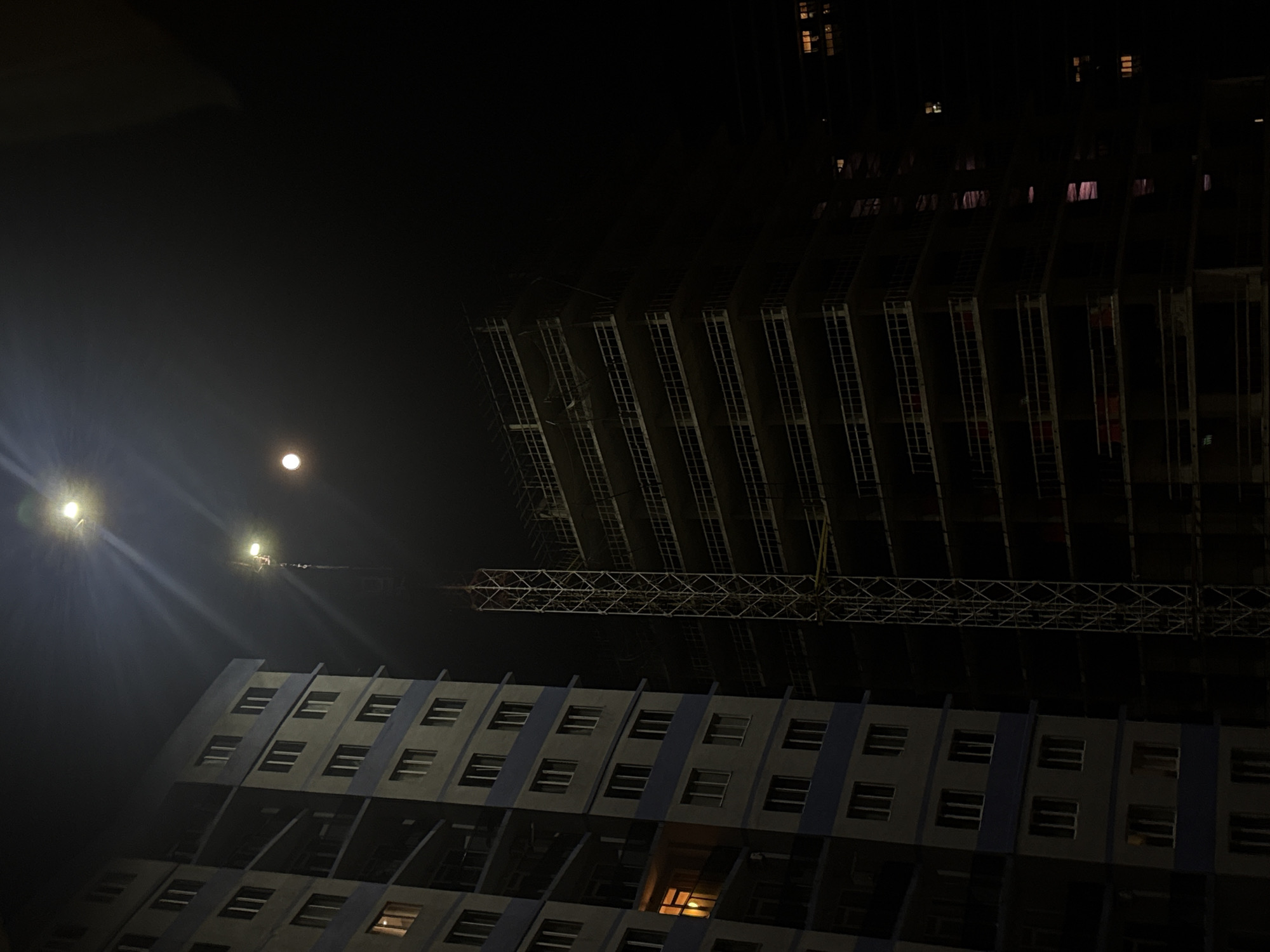 The image shows a night scene in Batumi, Georgia. In the foreground is a multi-story building with multiple rows of windows. Above it is a construction crane and an unfinished building, showing skeletal scaffolding. In the upper-left corner, there are two bright lights, and a faint moon can be seen.