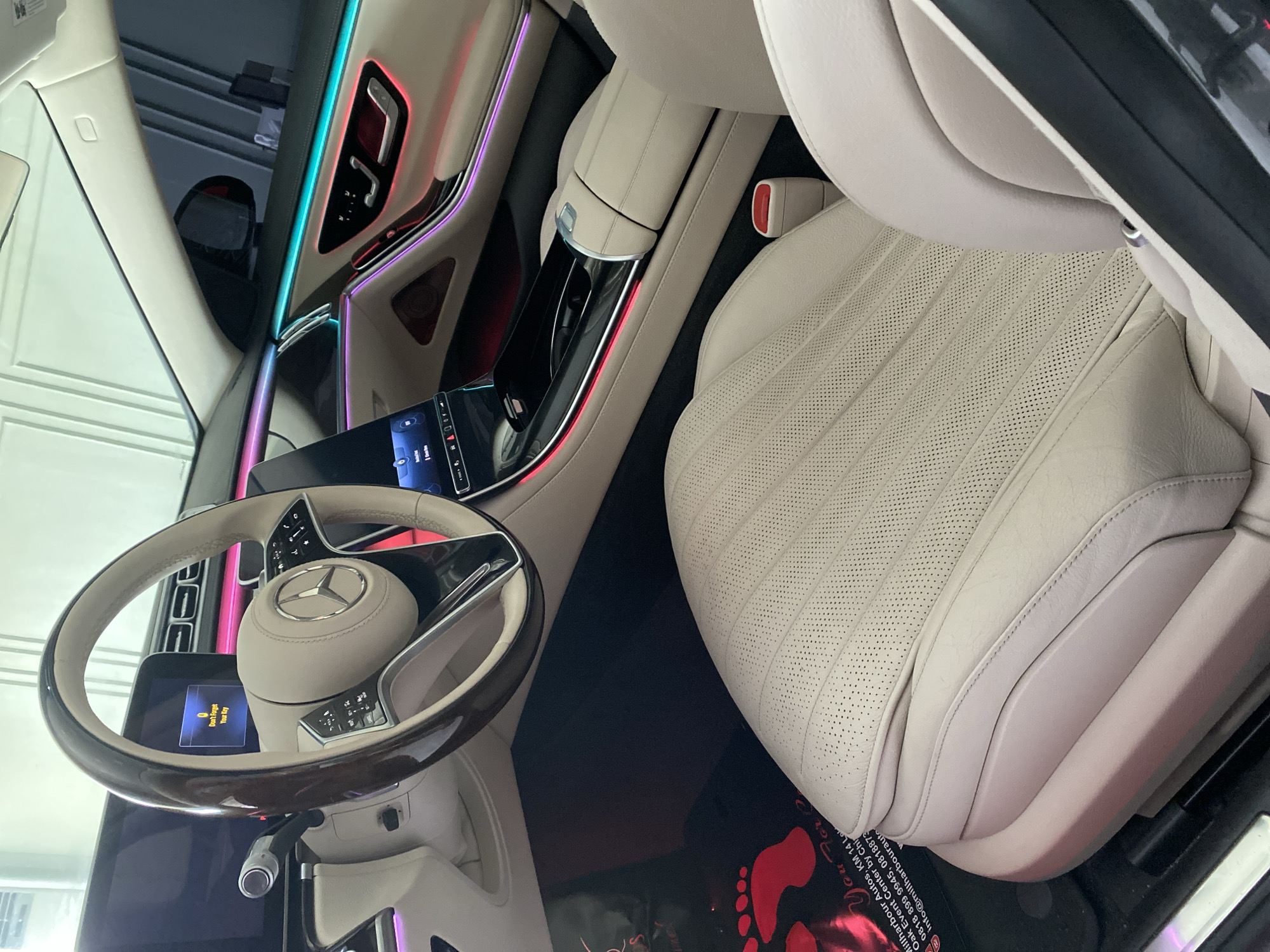 The image shows the interior of a luxury car, focusing on the front passenger seat and dashboard area. The seat is upholstered in light-colored perforated leather. The dashboard features ambient lighting in multiple colors. A Mercedes-Benz steering wheel is visible, displaying a message: "Don't Forget Your Key." On the floor of the car is a red-and-black floormat with the text "You Fore", followed by the name "Millharbour Autos, KM 14 Lok Oak Event Center by Chi". The location is therefore inferred to be Igboefon, Nigeria.