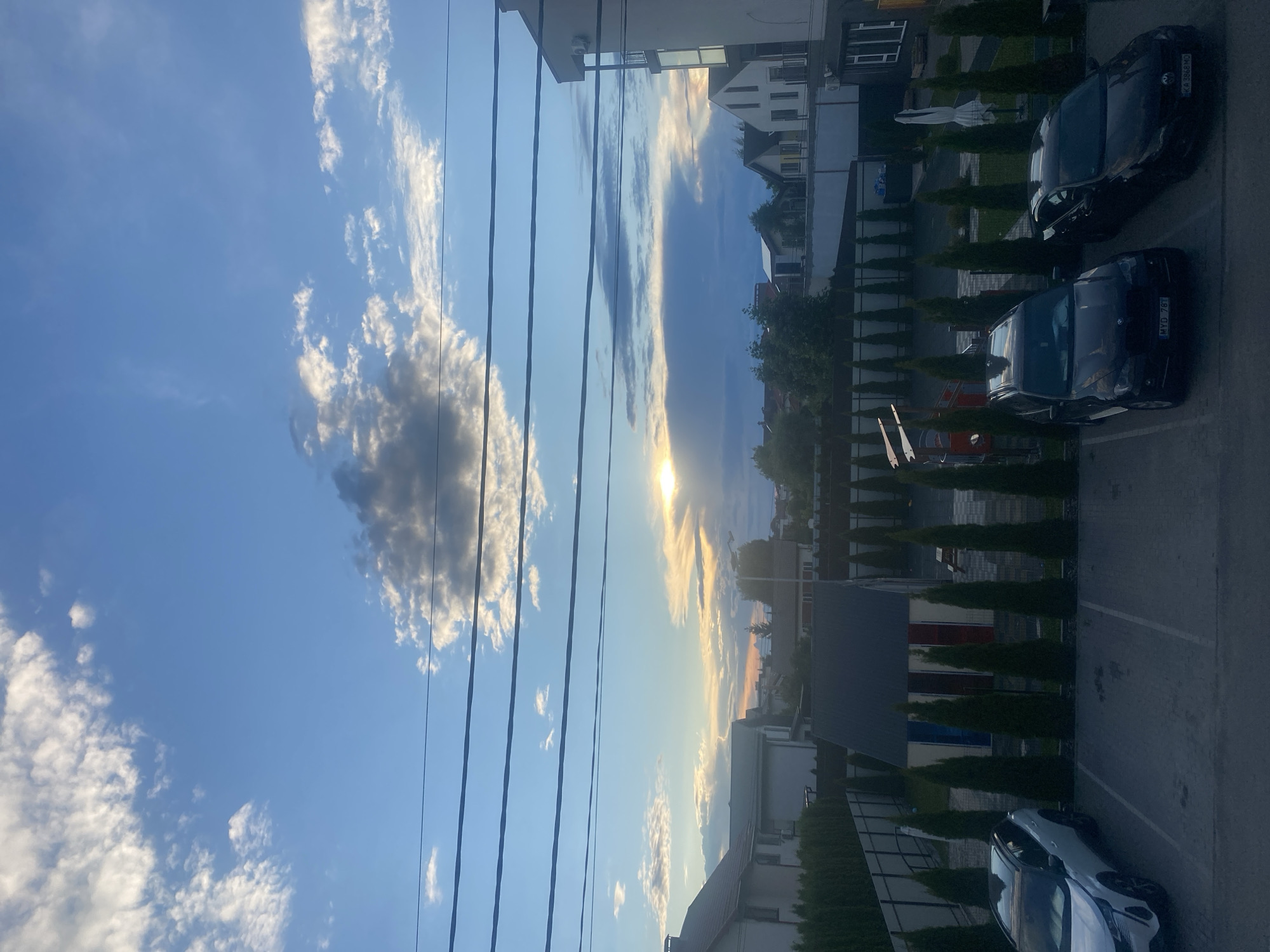 The image depicts a twilight scene in Novi Petrivtsi, Ukraine. The sky is a mix of blue and grey clouds, with the sun setting or rising behind a large cloud bank, casting a warm, golden light. Several parallel power lines stretch across the upper portion of the frame. Below the sky, a residential area is visible, characterized by houses with tiled roofs and a modern building with balconies. In the foreground, there is a paved parking lot with several vehicles. A white SUV is parked on the left, followed by two dark-colored cars. A row of manicured evergreen trees runs along the parking lot and separates it from a yard. Within the yard, there is a small building with colorful doors and a playground structure. The overall impression is a quiet suburban or semi-rural setting at dusk. There are no people visible in the image.