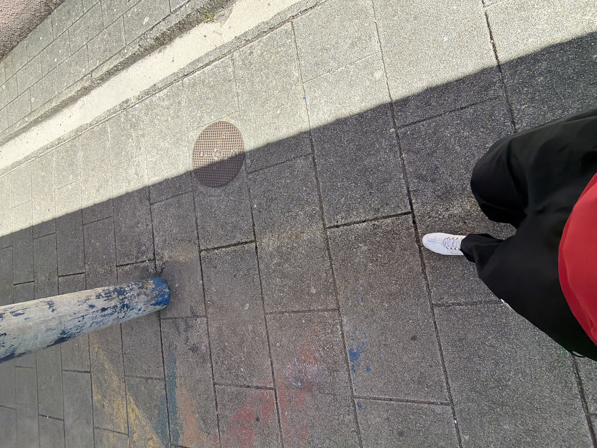 The image depicts a downward-angled, first-person view of a paved urban sidewalk in Vila Real, Portugal, under bright sunlight. The lower portion of an individual is visible in the bottom-center, wearing black trousers and a white athletic shoe on the right foot, with a red garment partially visible at the waistline. The pavement consists of rectangular grey concrete pavers, some displaying faded markings or graffiti in red, blue, and yellow. A weathered, cylindrical pole, predominantly grey with patches of faded blue paint, stands in the upper-left quadrant. A circular metal manhole cover with a grid pattern is positioned on the pavement towards the mid-right. A strong diagonal shadow line bisects the scene, indicating a direct light source from the upper right, illuminating the right side of the pavement and leaving the left side in shade. Along the far right edge, a narrow concrete drainage channel runs parallel to a textured wall or curb.